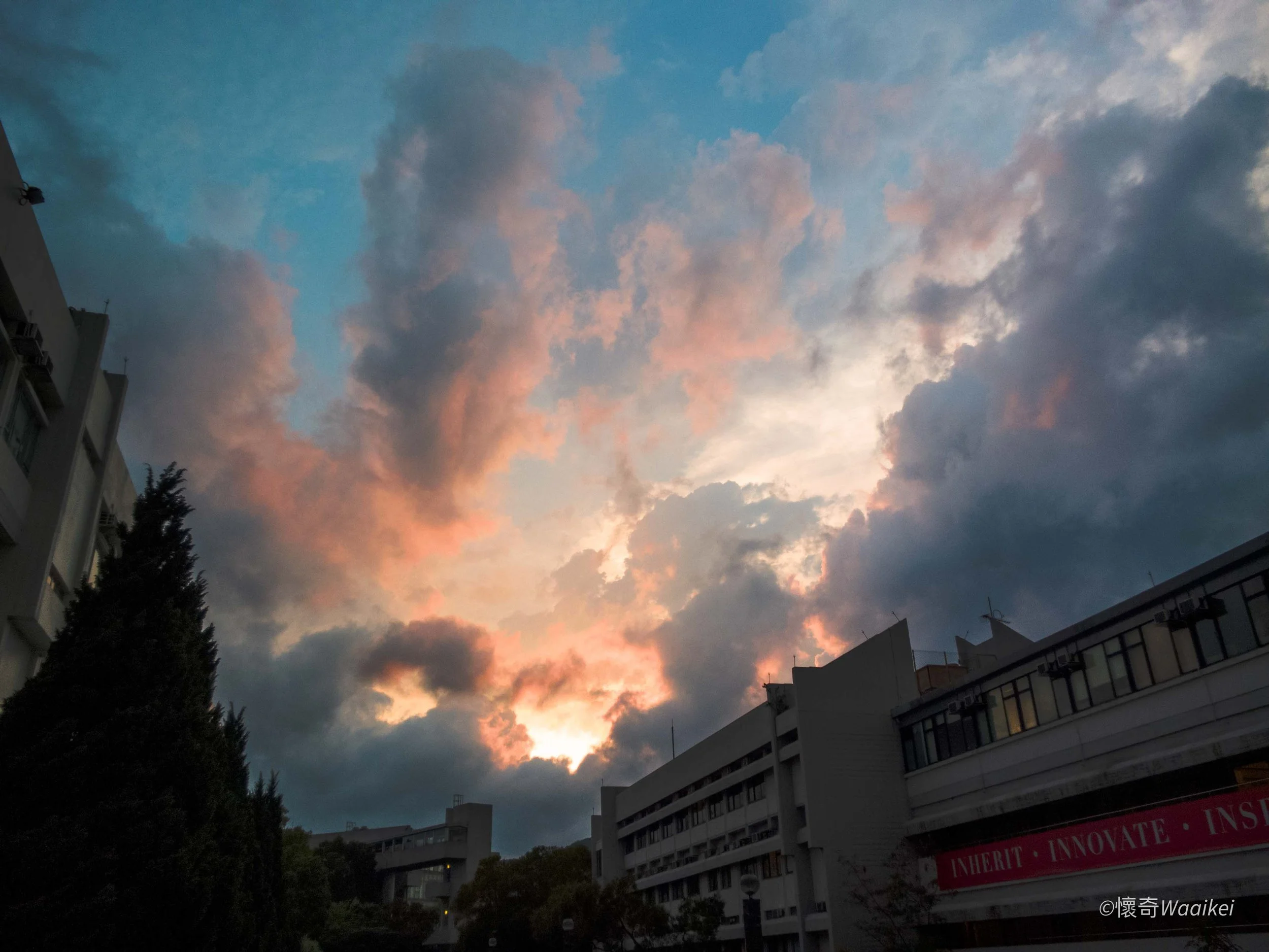 Sky with clouds at sunset or sunrise over urban buildings and trees.