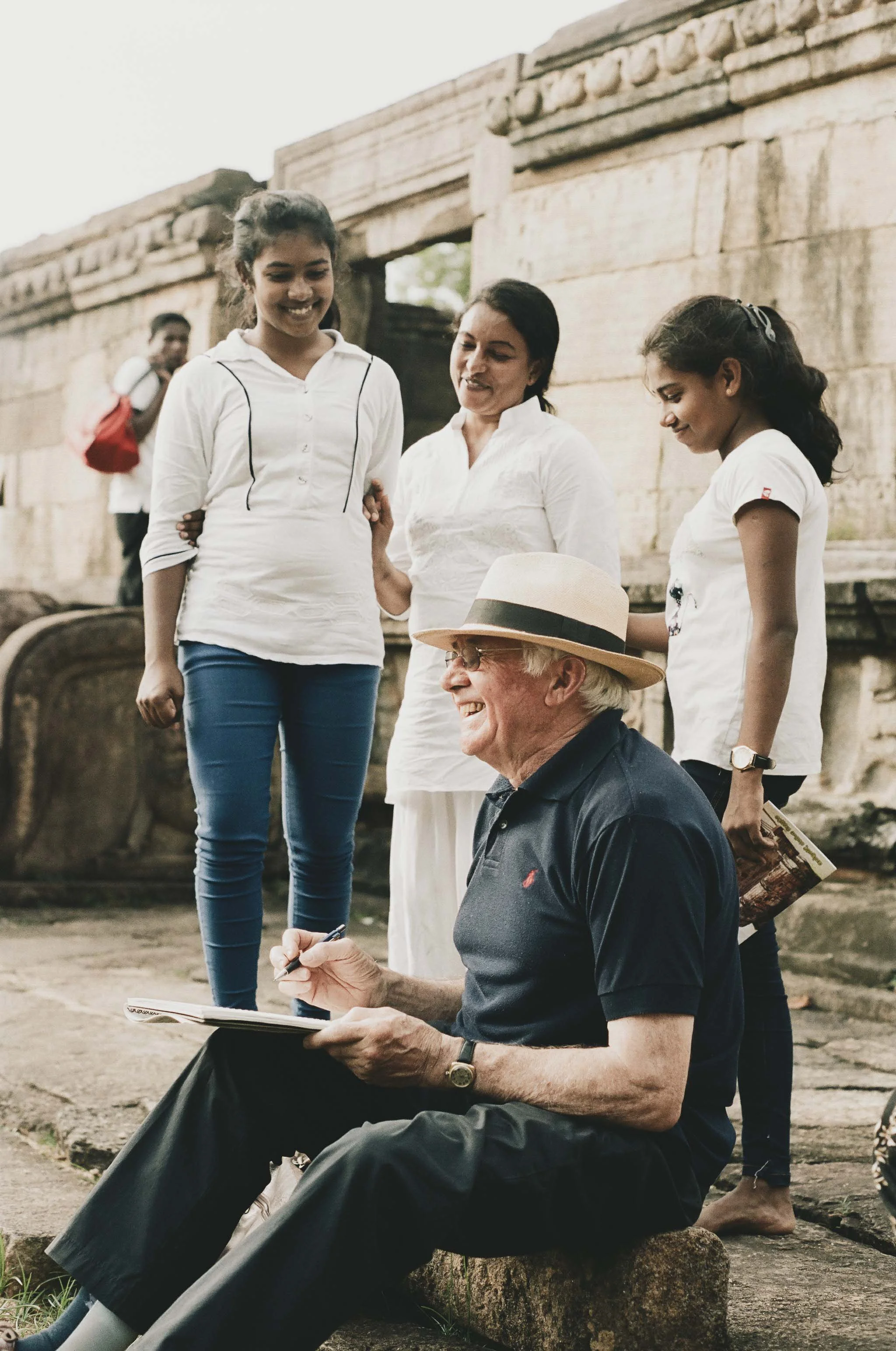 A group of four people, three young girls and an elderly man, gathered outdoors near a stone structure, with the man sitting on a rock and signing a book, while the girls and another woman stand nearby, smiling and engaging in conversation.