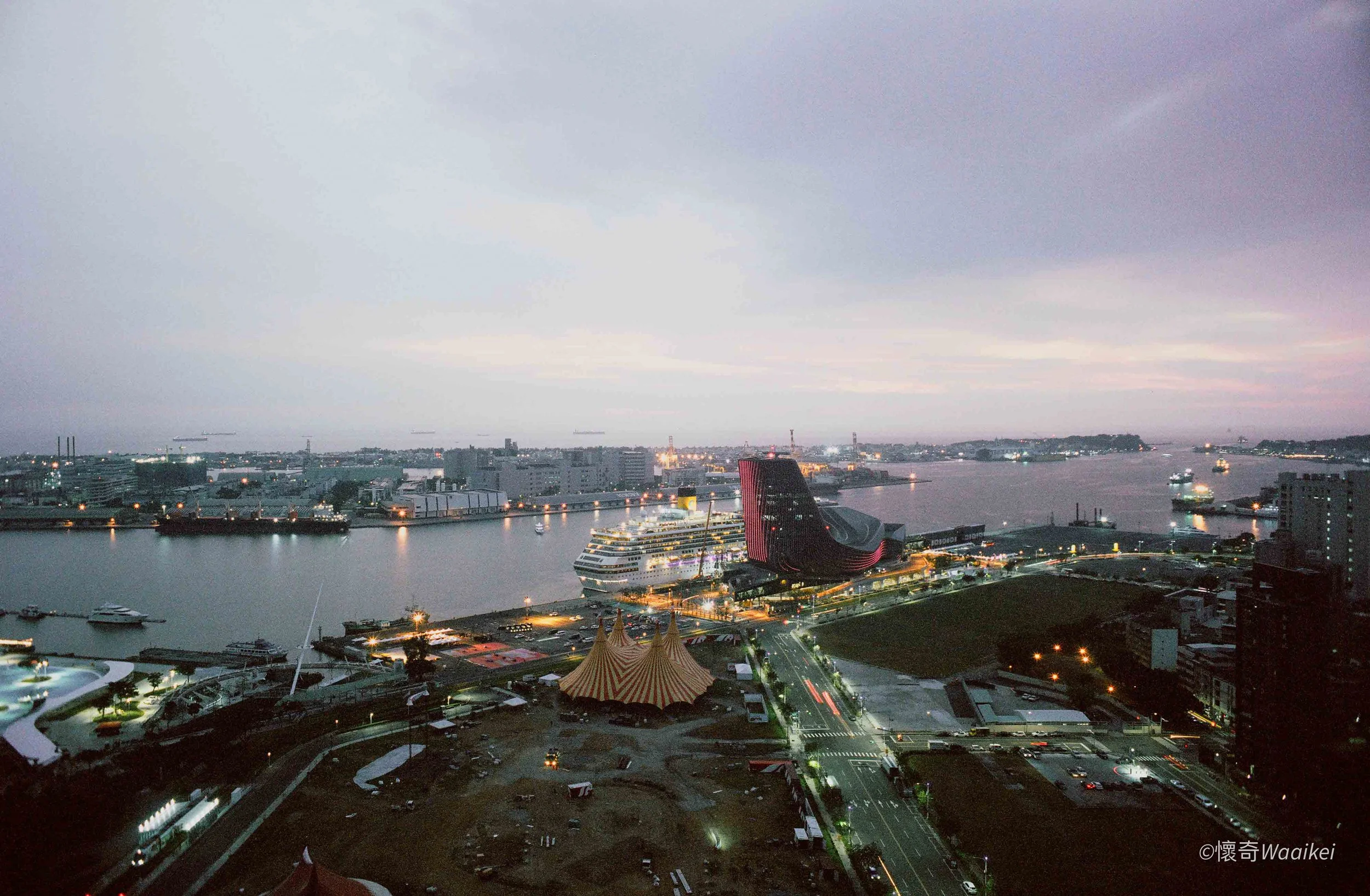 City skyline at dusk with a river in the foreground, cruise ship docked at a port, and cloudy sky overhead.
