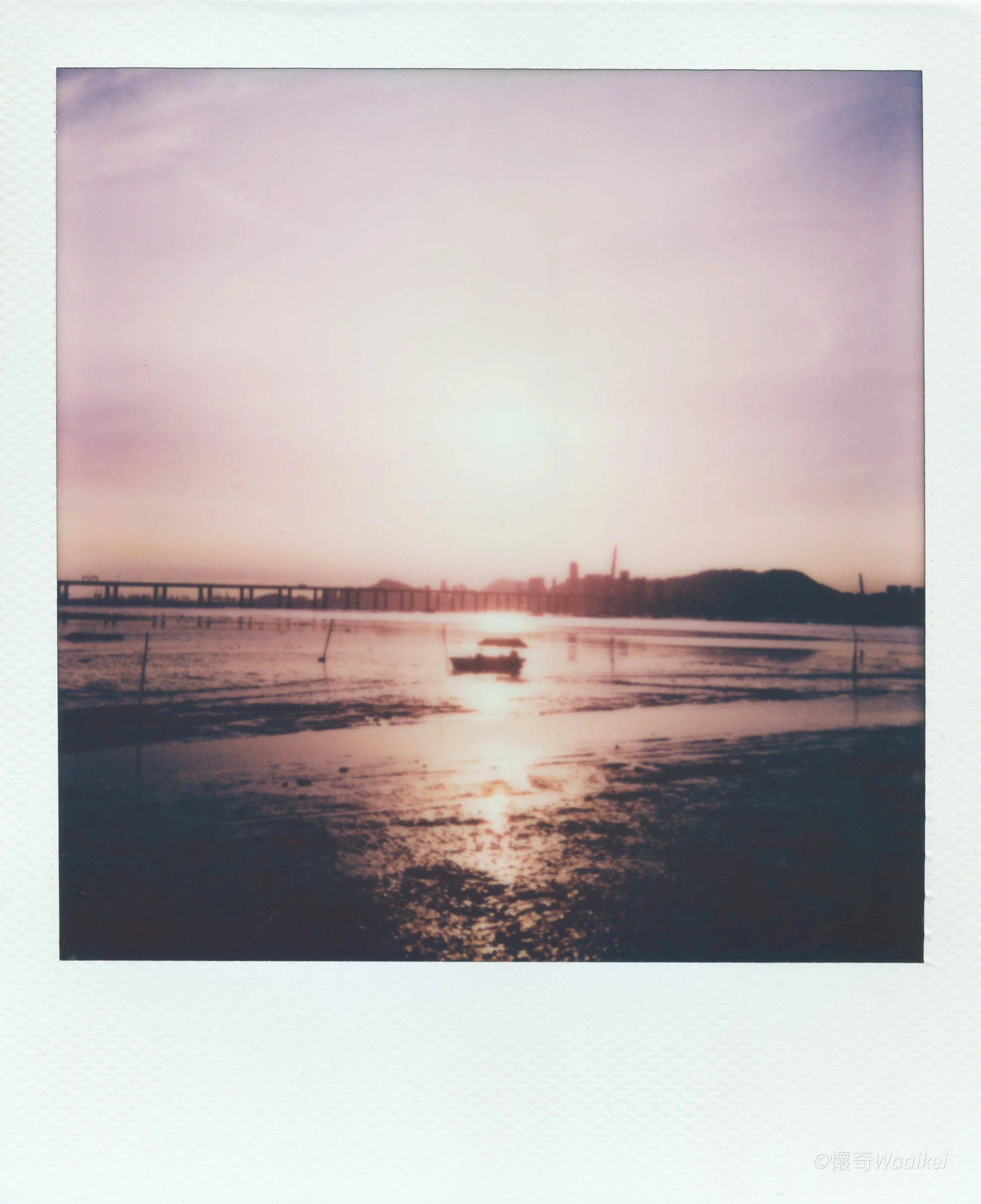 Sunset over a body of water with a silhouette of a boat and a bridge, with purple and orange sky.