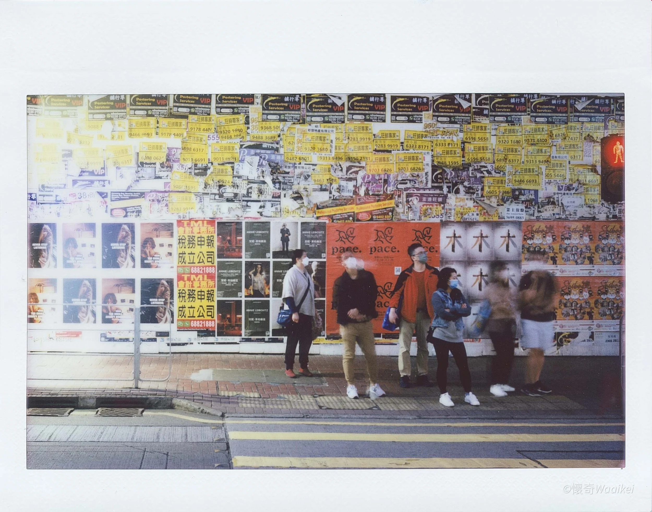 People waiting at a bus stop in front of a wall covered with yellow and black advertisements.