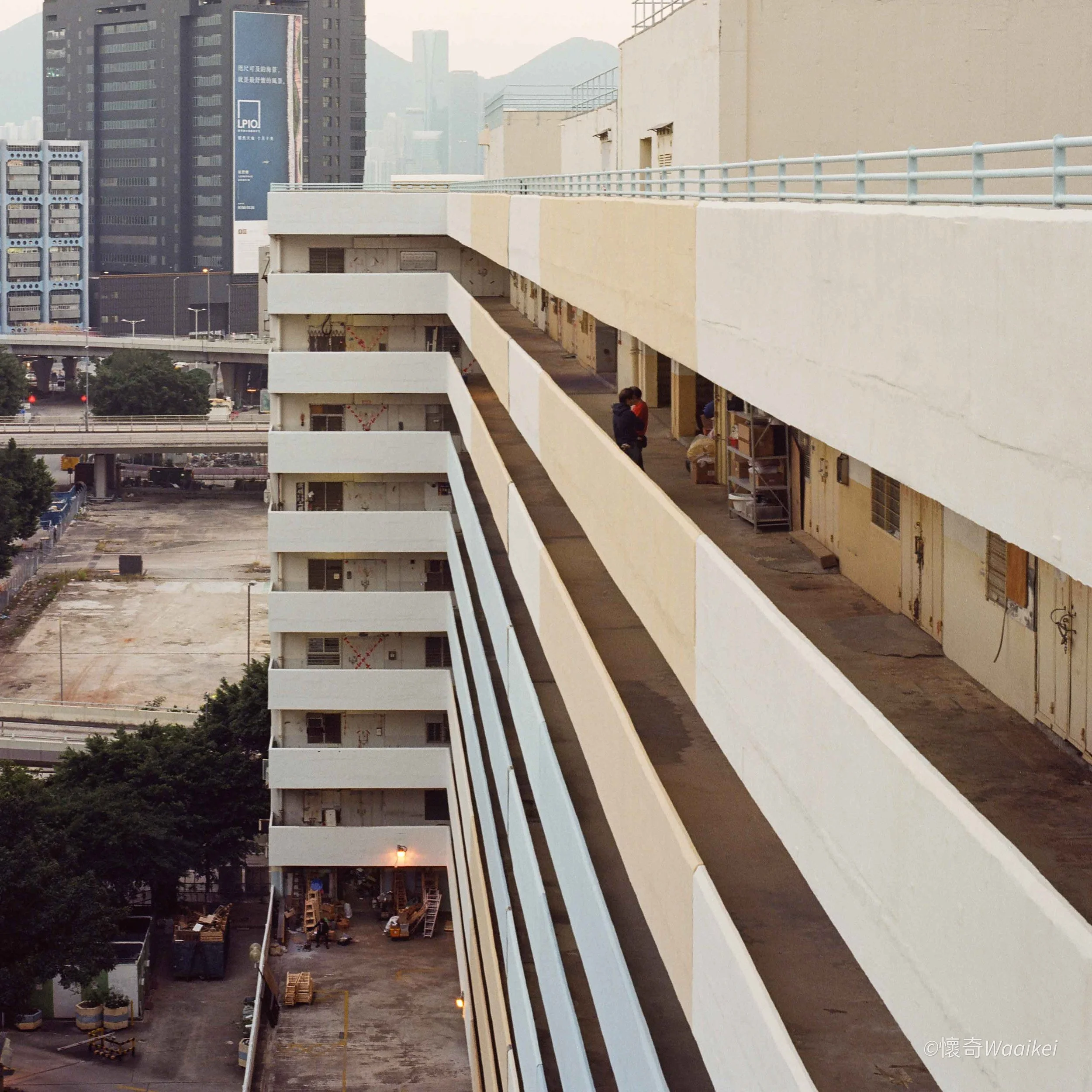 Construction site with unfinished multi-story building, workers, and equipment in an urban area with tall buildings in the background.