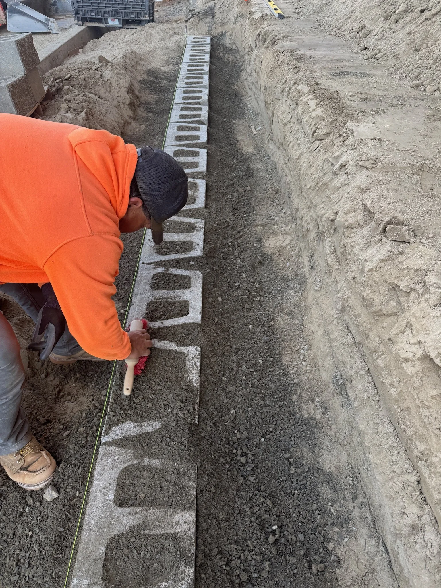 A construction worker wearing an orange jacket, black cap, and protective boots is laying concrete blocks along a narrow trench. The worker is using a tool to position the blocks, and a string line runs parallel to the trench to ensure straight alignment. The dirt around the trench is dug out, and construction equipment is visible in the background.