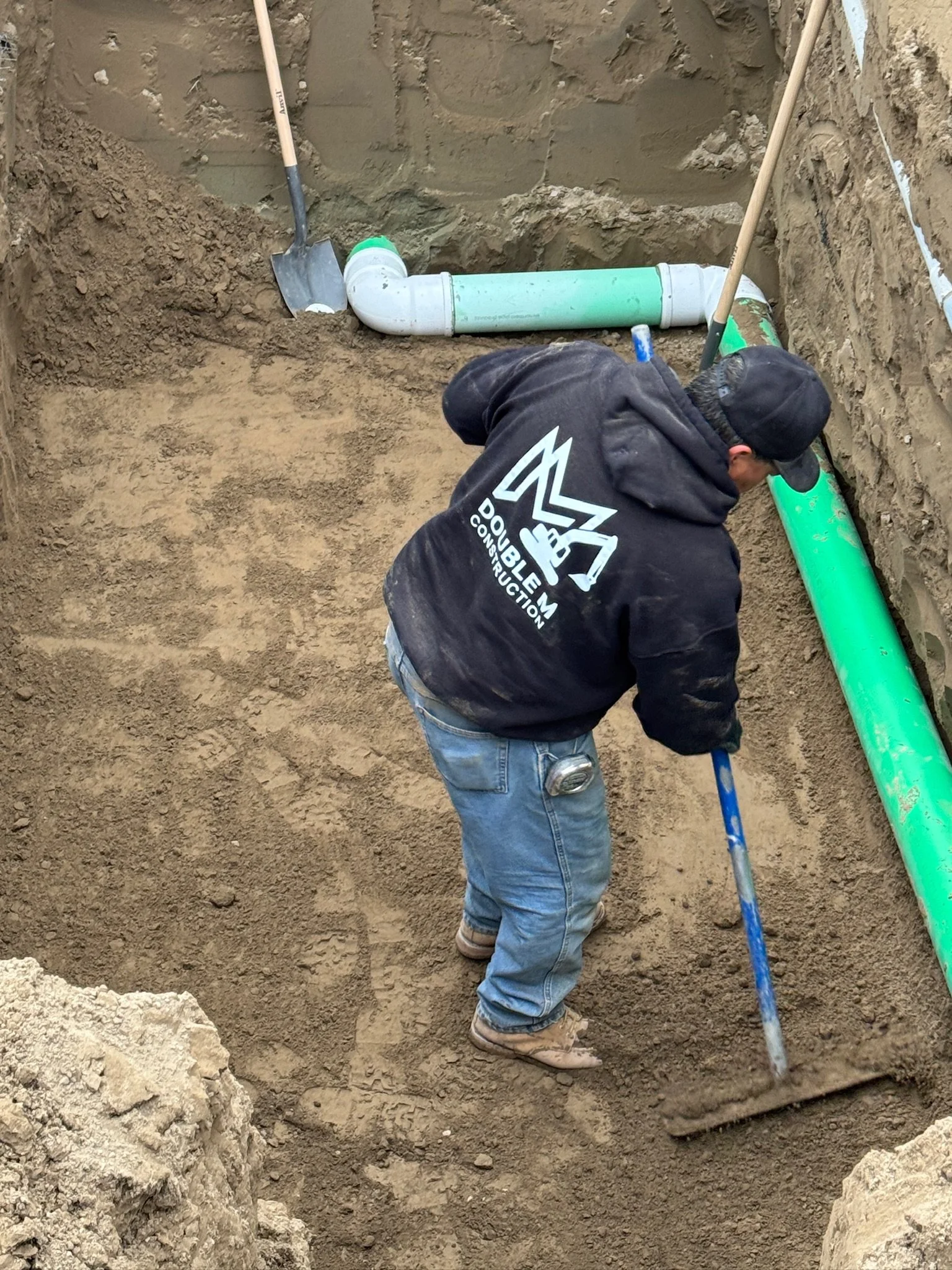 A construction worker examining underground plumbing pipes in a dirt trench. The worker wears a black hoodie with construction company branding, jeans, and boots, and holds a measuring stick. There are vertical shovels and a green pipe in the trench.