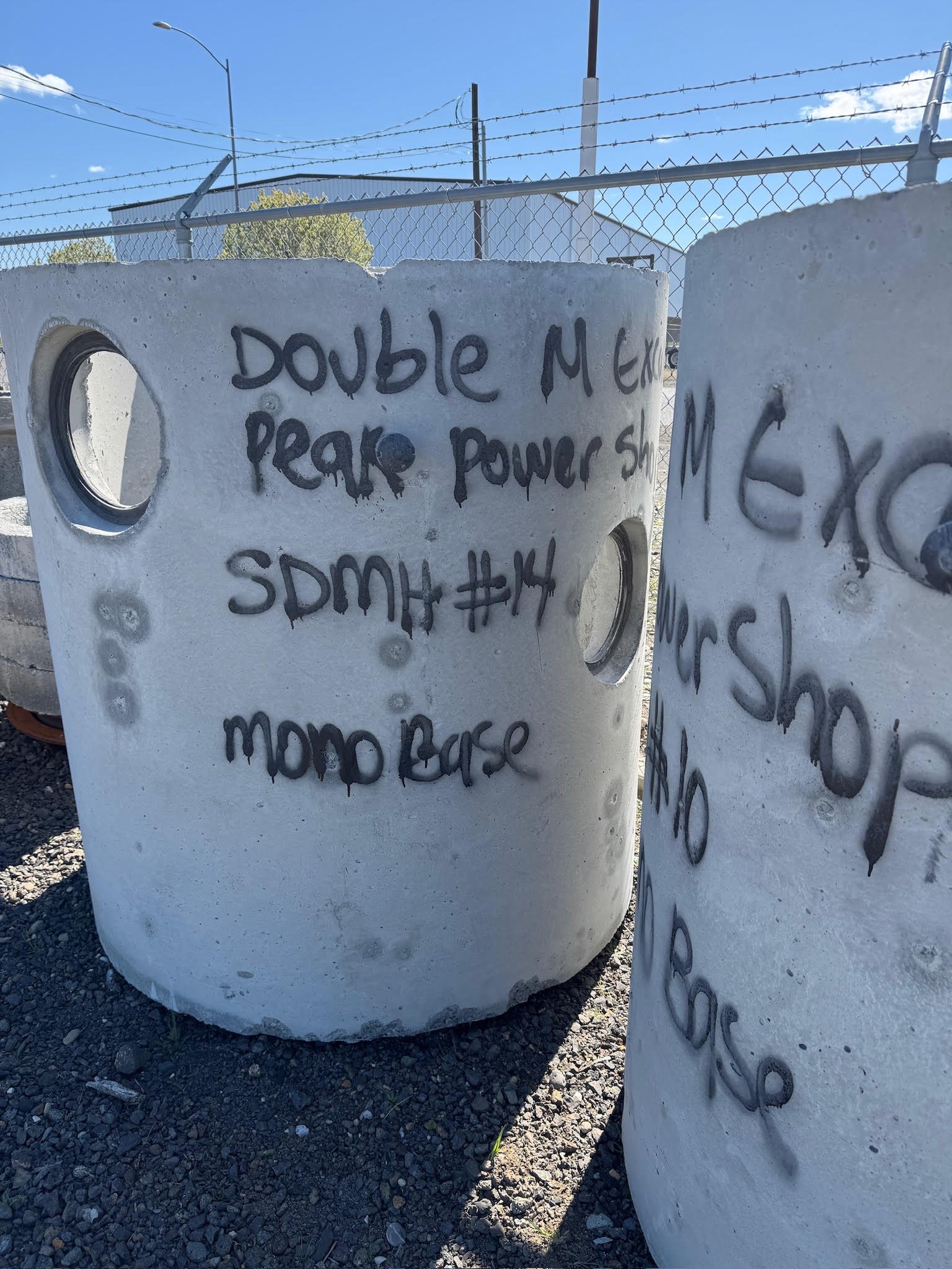 Concrete blocks with spray-painted messages and symbols, situated outdoors on gravel, with a chain-link fence and blue sky in the background.