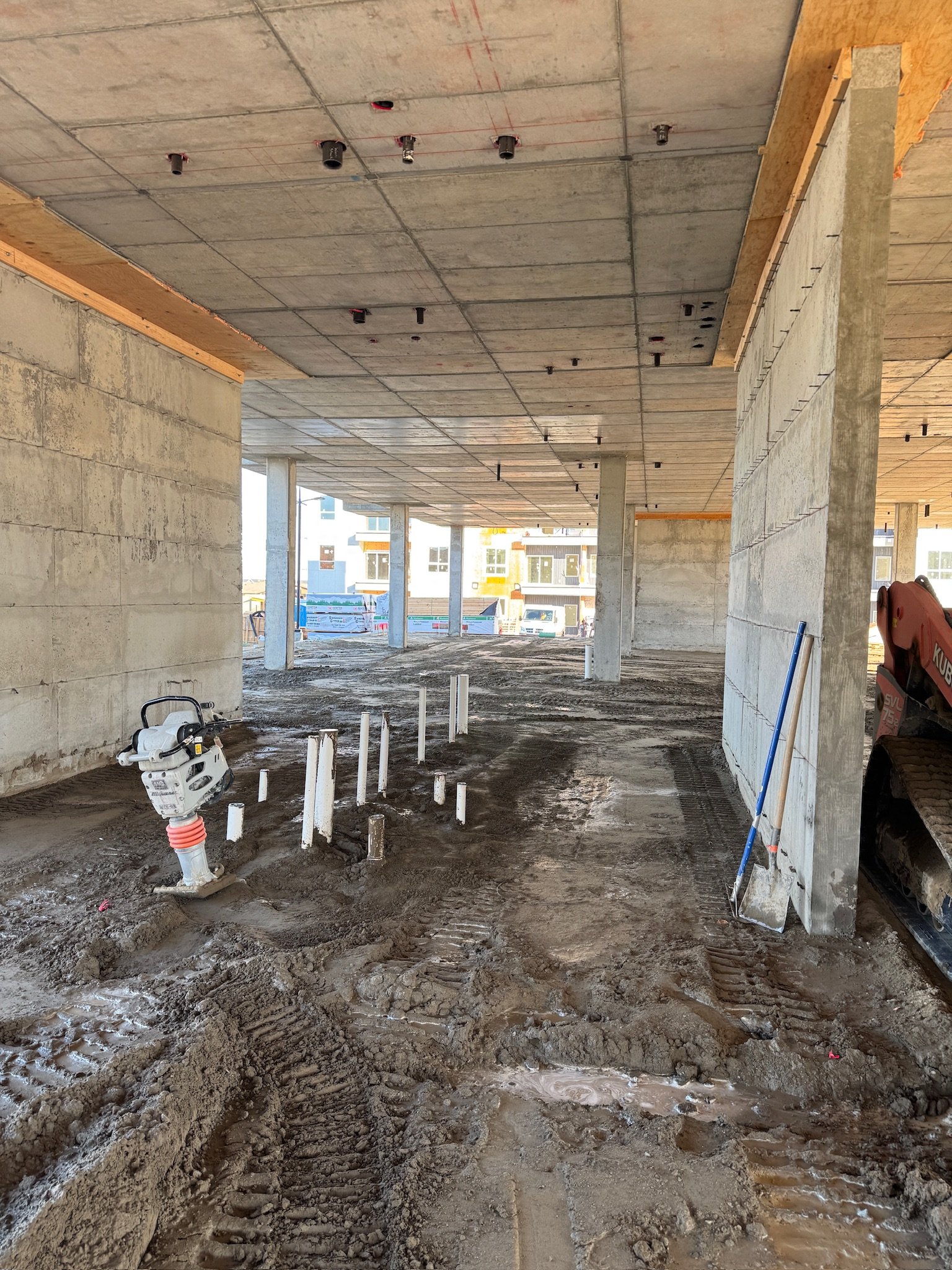 Construction site of a building with concrete columns and unfinished flooring. Construction equipment and pipes are visible, with piles of dirt and tire tracks on the ground.