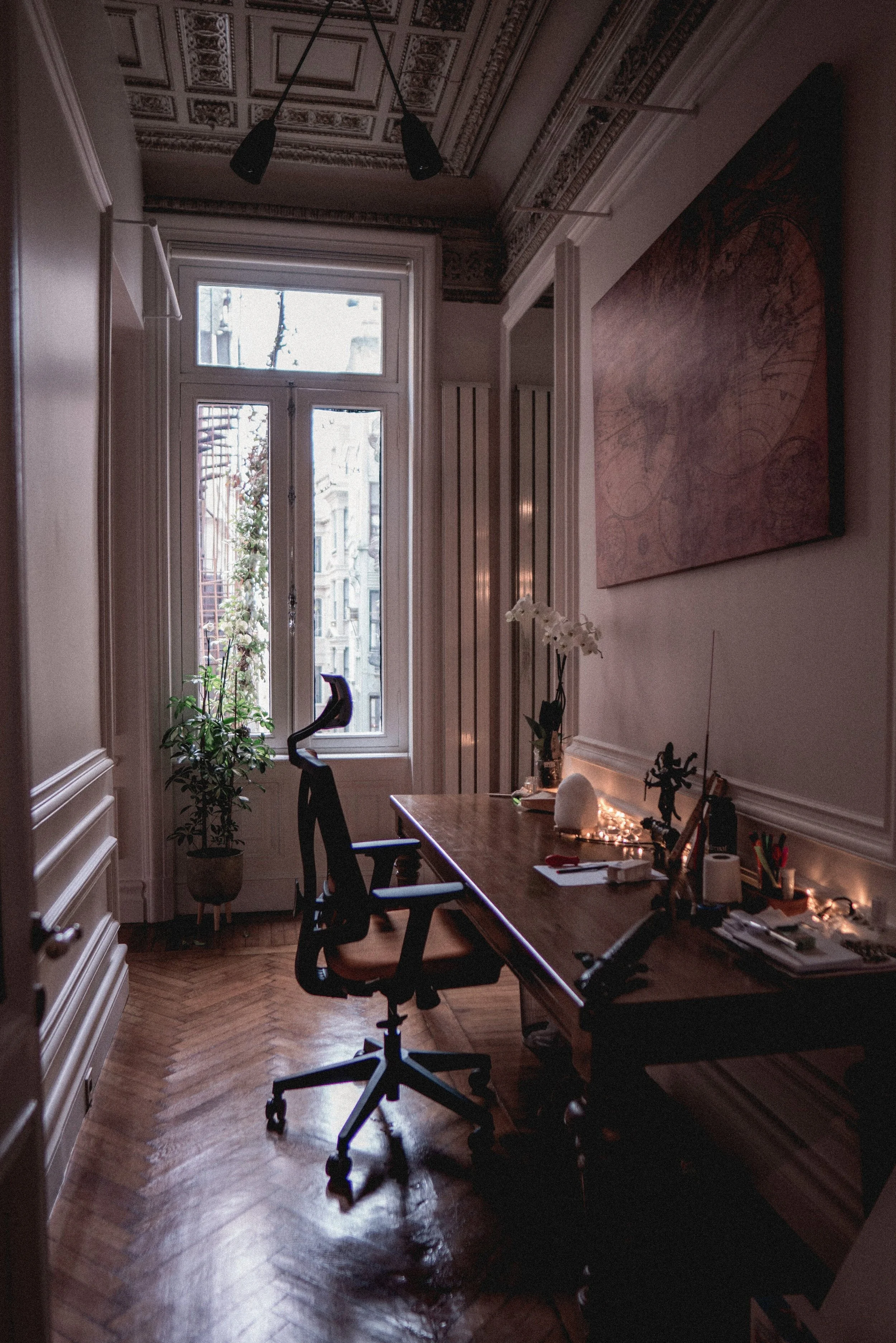 An office with a large wooden desk, two office chairs, a window with city buildings outside, a potted plant, a painting on the wall, and various office supplies and decorations.