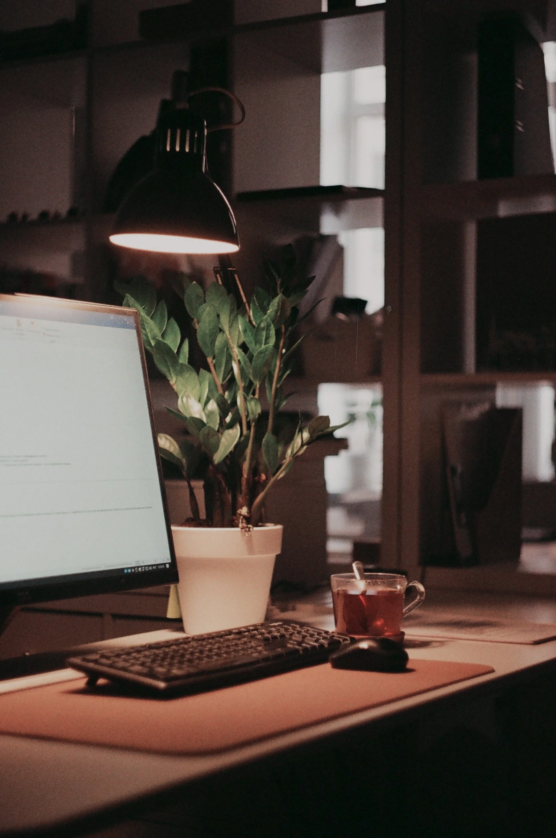 A computer desk with a monitor, keyboard, mouse, a potted plant, a glass cup filled with tea, and a black lamp in a dimly lit room with wall shelves in the background.