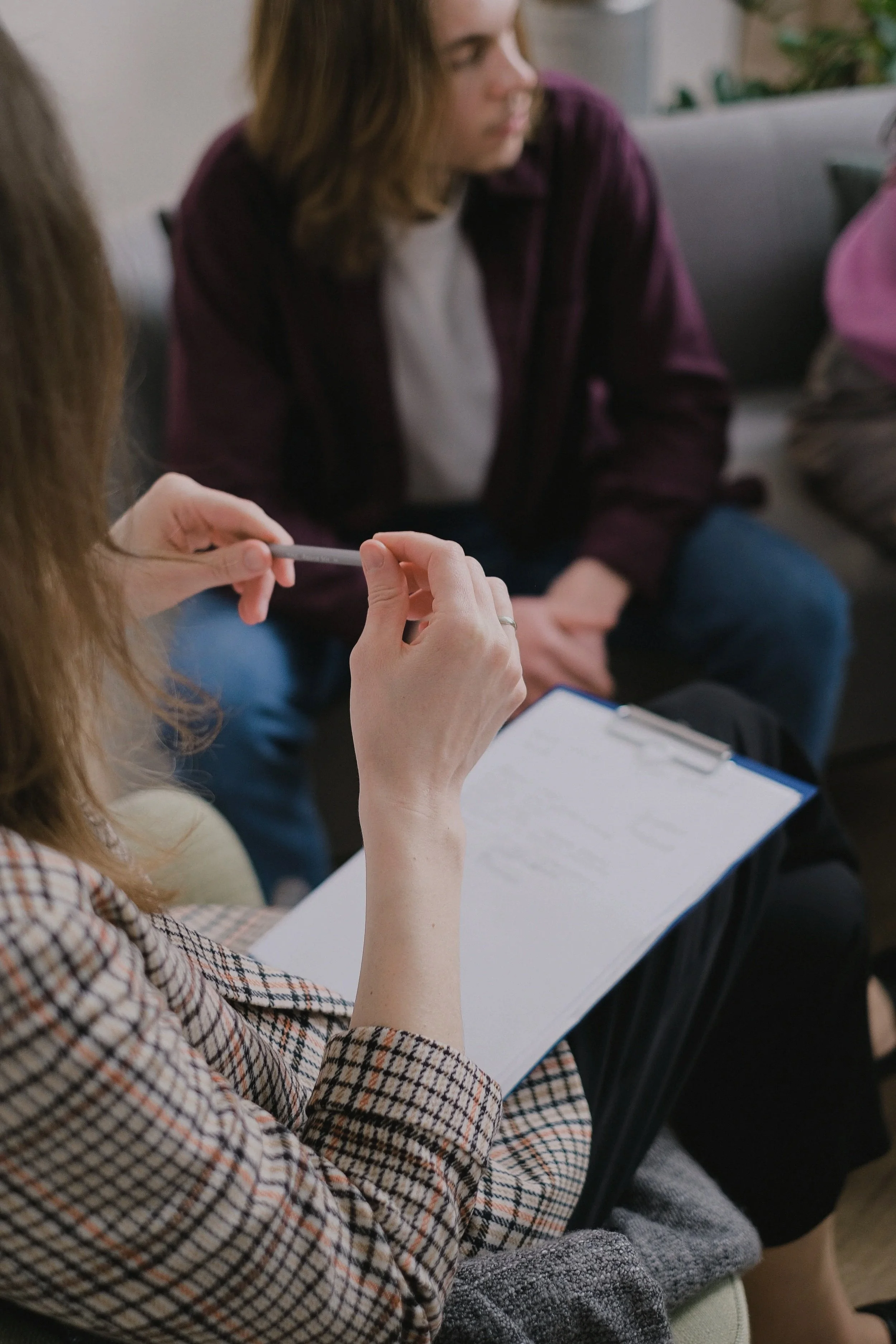 A woman with long hair wearing a checkered blazer sitting with other people during a meeting or class, holding a pen and a notebook.