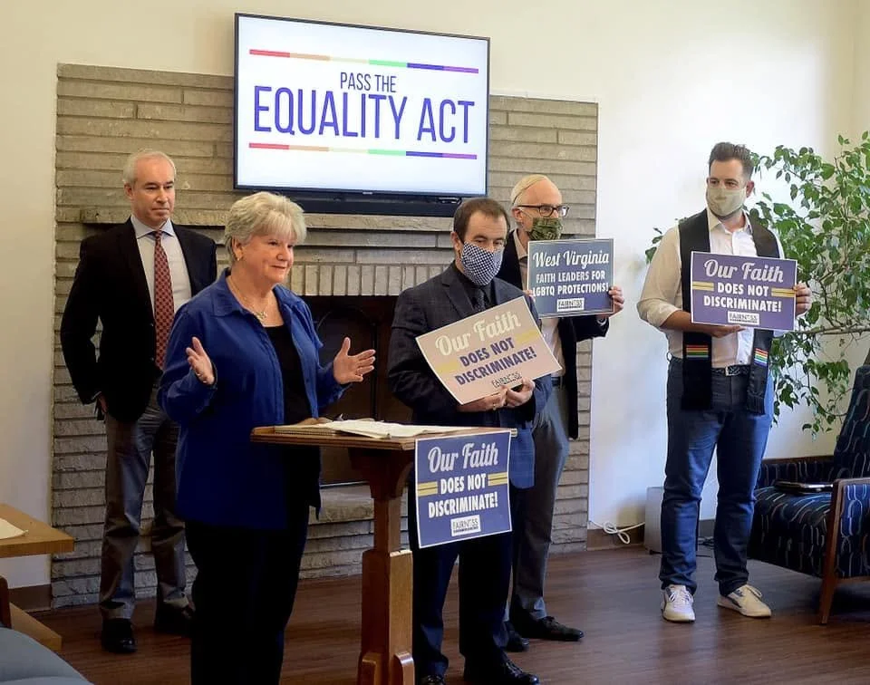 Group of five people speaking at a rally or press conference, with a large screen displaying 'PASS THE EQUALITY ACT,' and holding signs that read 'Our Faith Does Not Discriminate.'