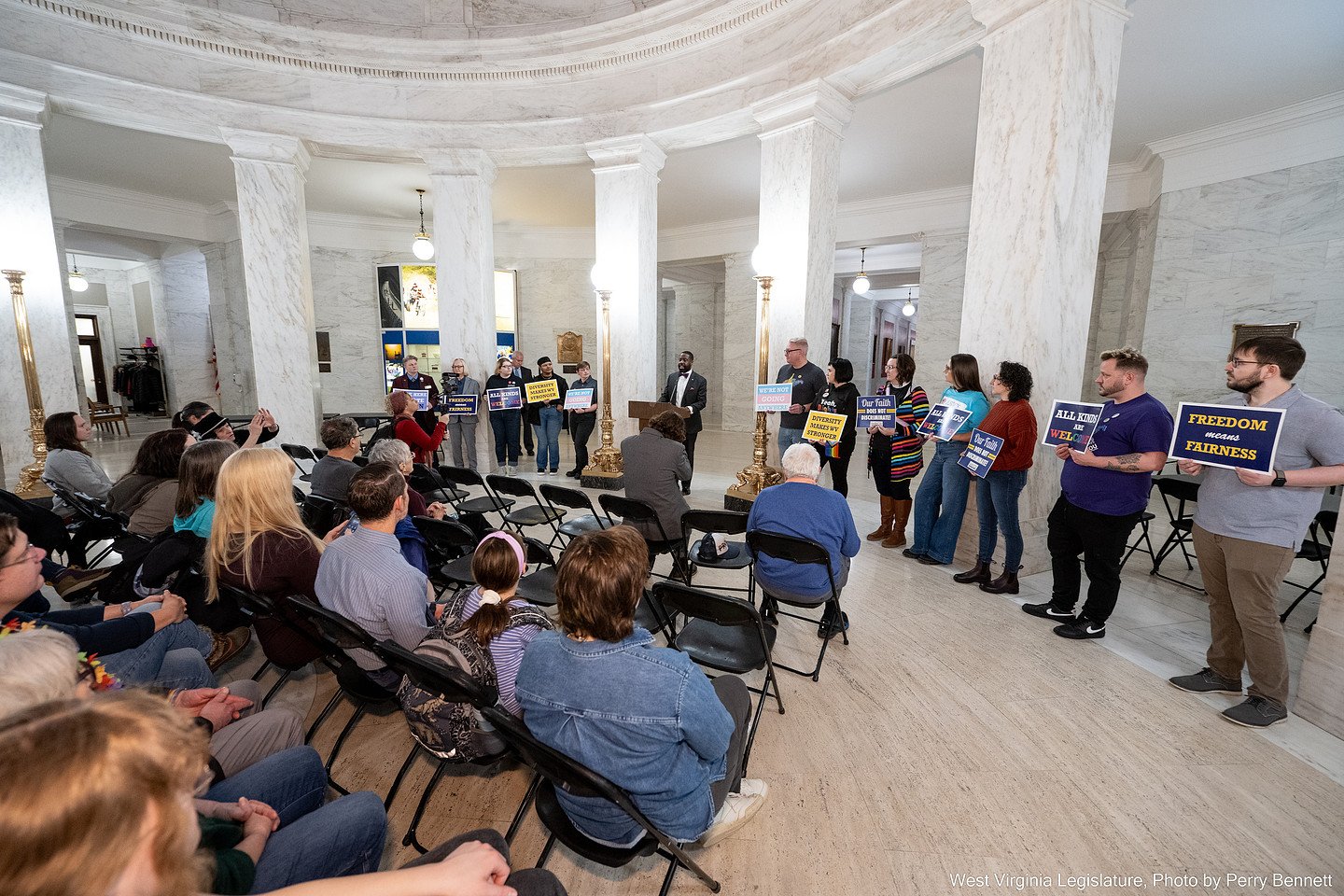 People holding signs at a rally inside a building with marble columns and floors, addressing an audience seated in chairs.