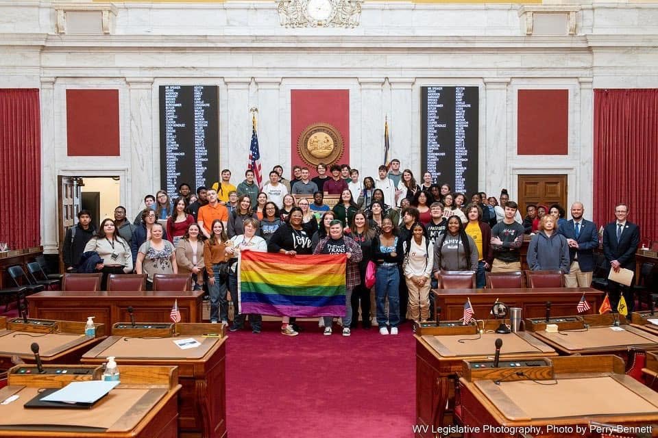 Group of diverse people holding a rainbow pride flag inside a government chamber, with official seating and large display boards in the background.