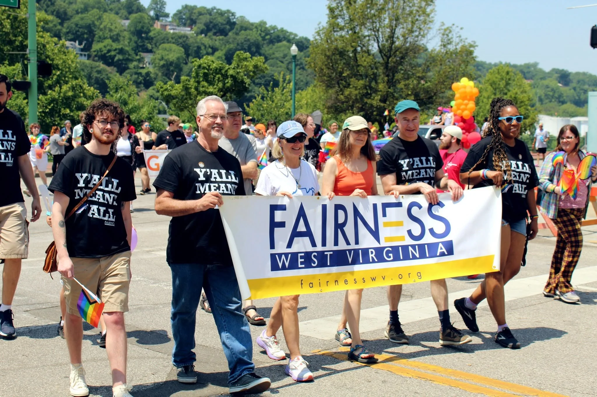 Group of people participating in a pride parade holding a banner that reads 'Fairness West Virginia.' Several attendees wear shirts with slogans, some are wearing rainbow accessories, and there are colorful balloons in the background.