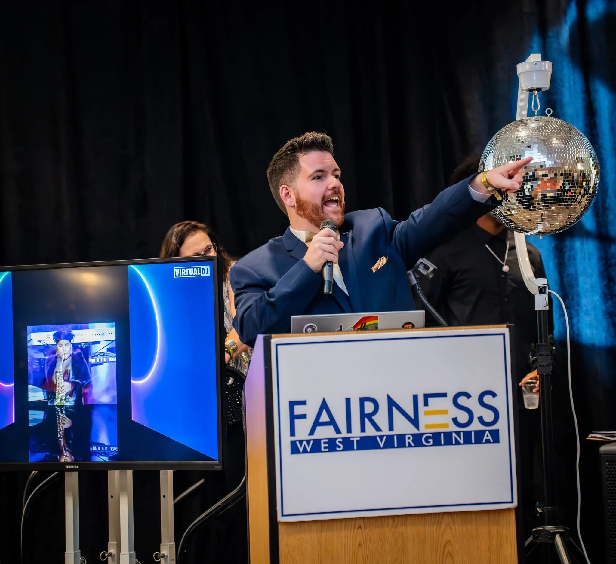 Man with a beard and suit gesturing toward a disco ball at an event, with a woman wearing glasses nearby, monitor screen displaying a DJ logo, and a sign reading 'FAIRNESS WEST VIRGINIA' in the background.
