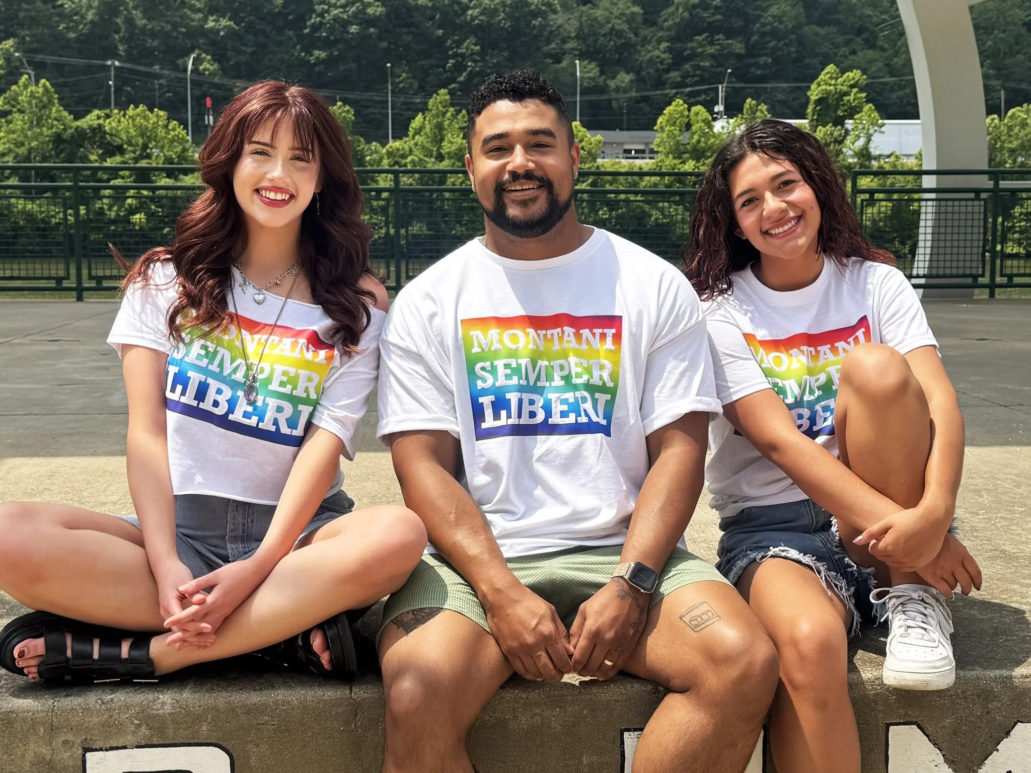 Group of three young adults sitting together outdoors on a sunny day, wearing white T-shirts with rainbow-colored text that reads "Montani Semper Liberi," with green trees and a fence in the background.