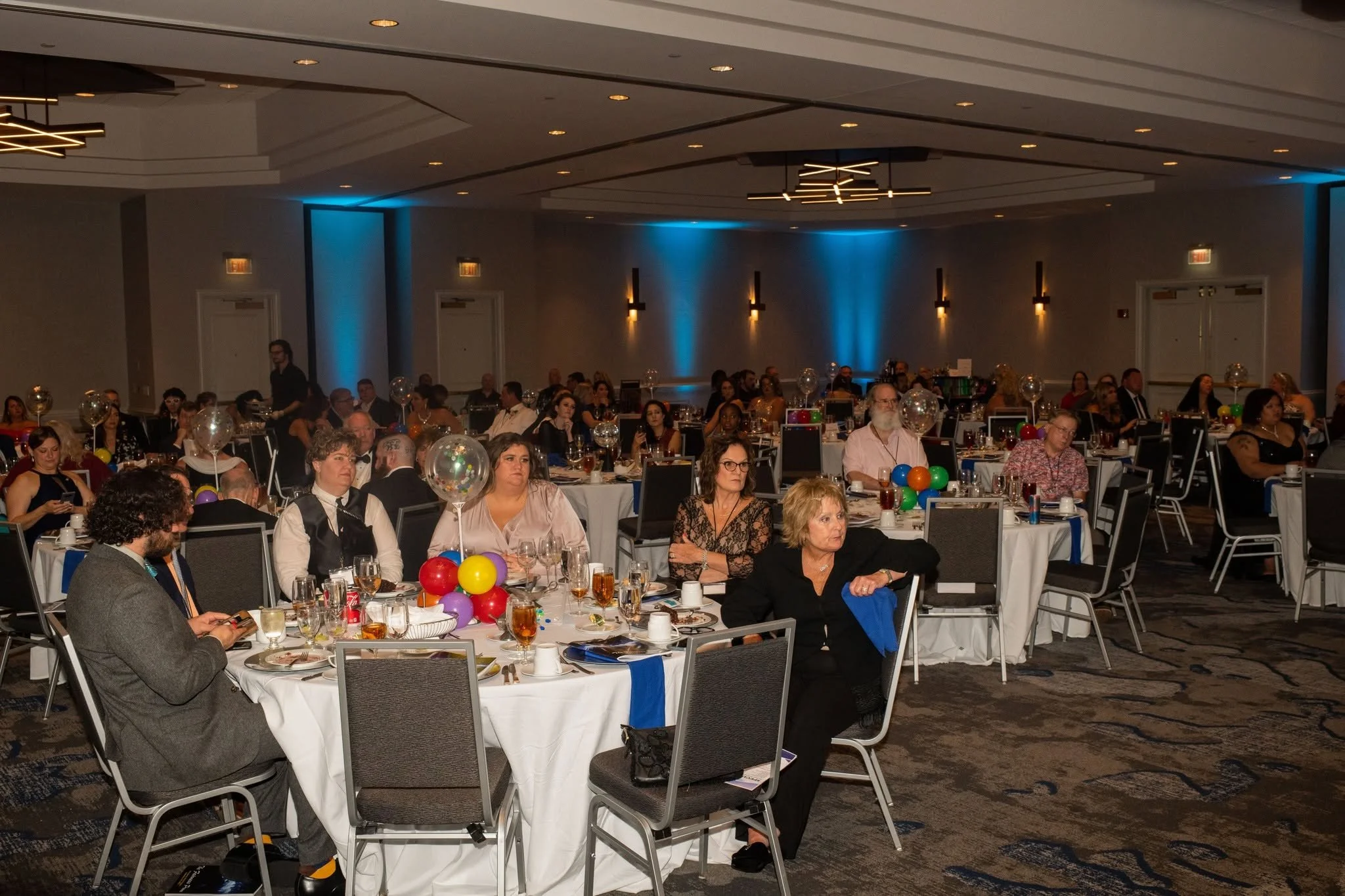 Indoor banquet hall filled with guests seated at round tables decorated with balloons. People are dressed formally, some are eating, talking, or using their phones. The room has modern lighting and blue accent lighting on the walls.