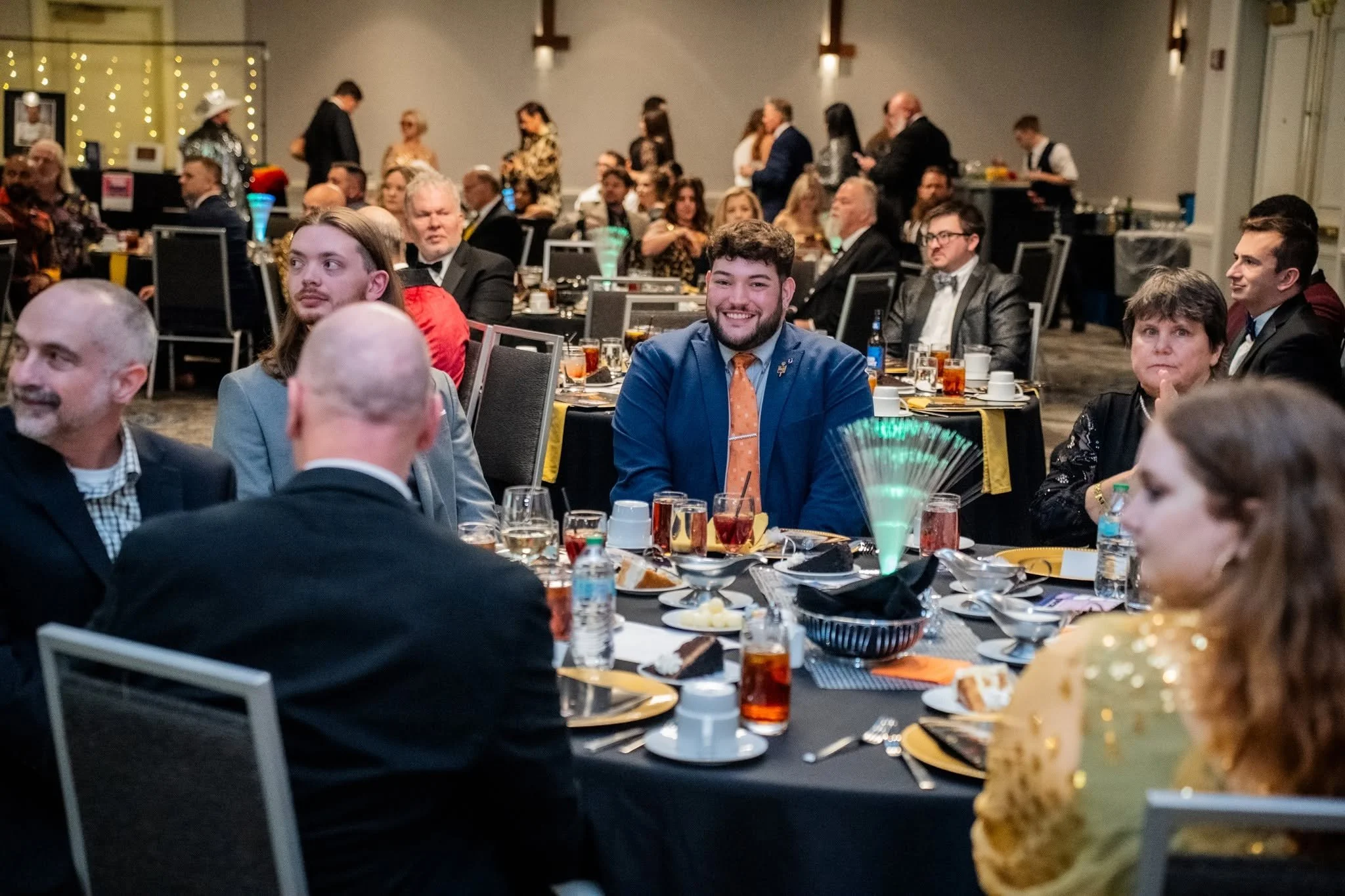 People attending a formal event or dinner, seated at round tables with black tablecloths, set with plates, glasses, and drinks, in a decorated banquet hall with string lights and wall decorations.