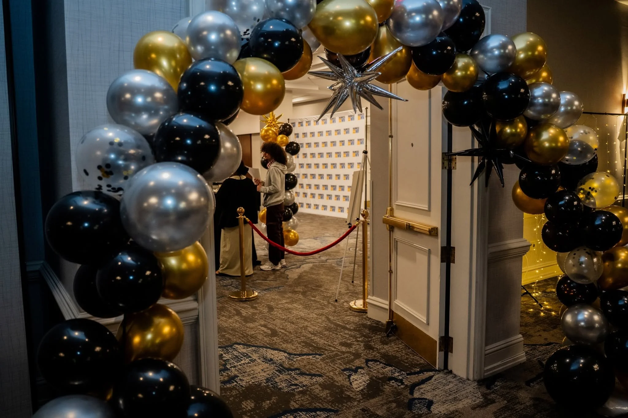 Entrance decorated with black, gold, and silver balloons forming an arch, leading into an event space with a backdrop and people standing inside.