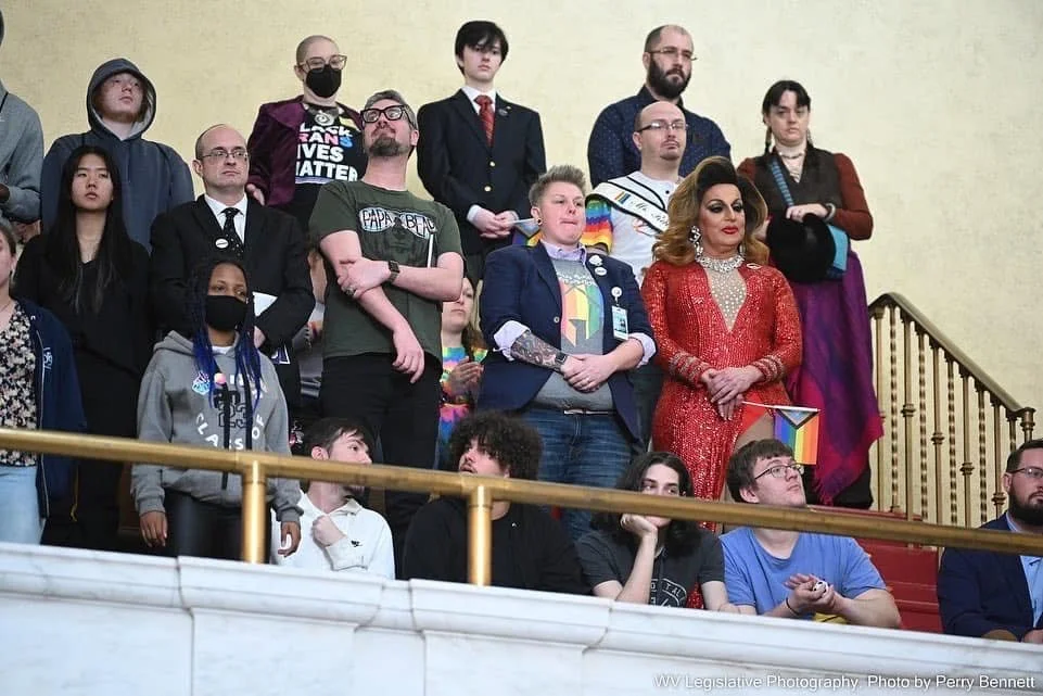 Group of diverse individuals standing and sitting on a staircase during a formal event, some wearing rainbow-colored accessories and holding LGBTQ+ flags, with a woman in a red dress and a rainbow-themed bag.