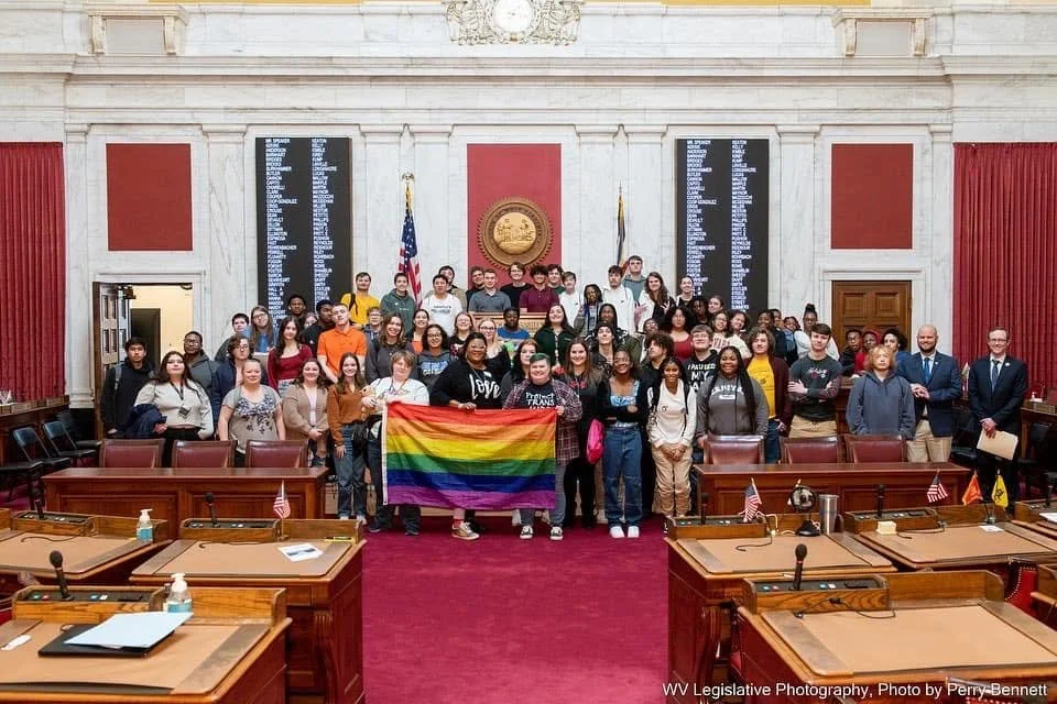 Group of diverse people inside a government chamber, some holding a rainbow pride flag, posing for a photo.