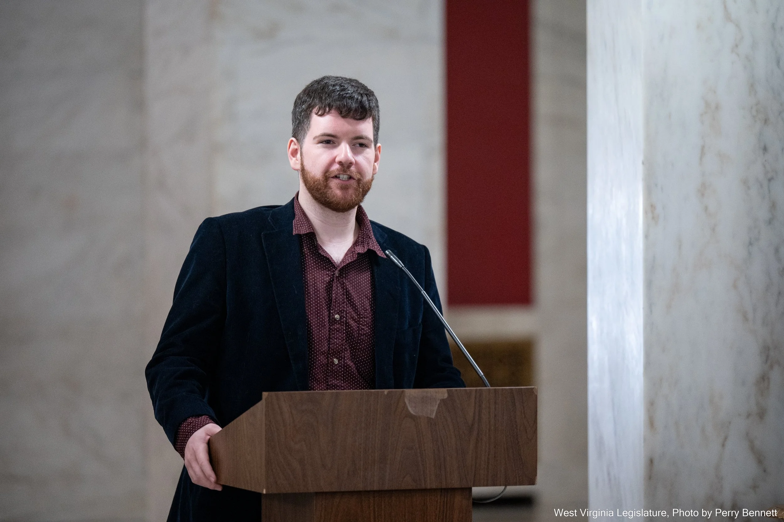 A man with a beard and mustache stands behind a wooden podium, speaking or giving a presentation in a formal setting. He is wearing a dark blazer and maroon shirt.