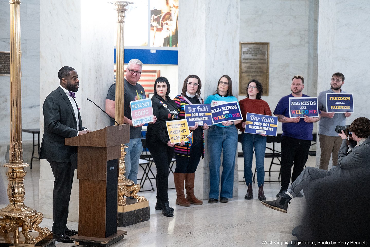 A man in a black suit and silver hair is speaking at a podium in a marble hall, while seven people stand behind him holding signs advocating for equality, acceptance, and fair treatment.