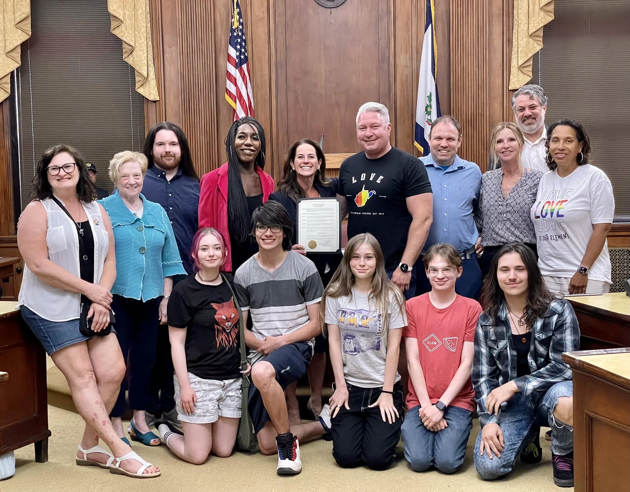 Group of diverse people, including children and adults, posing together inside a room with wood-paneled walls, American flags, and a framed document, suggesting a celebratory or official event.
