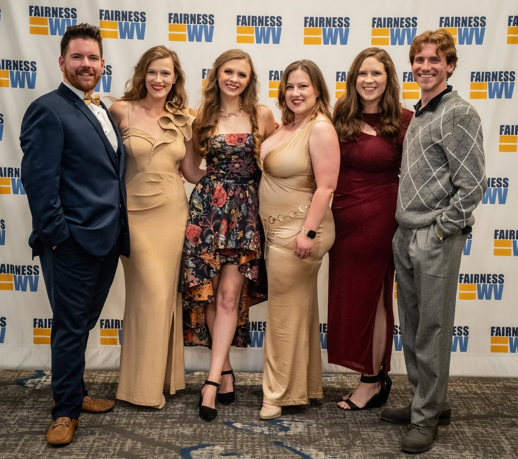 A group of six people, three men and three women, standing together and smiling at an event with a 'Fairness WV' backdrop behind them. They are dressed in formal attire, with the women wearing dresses and the men wearing suits.