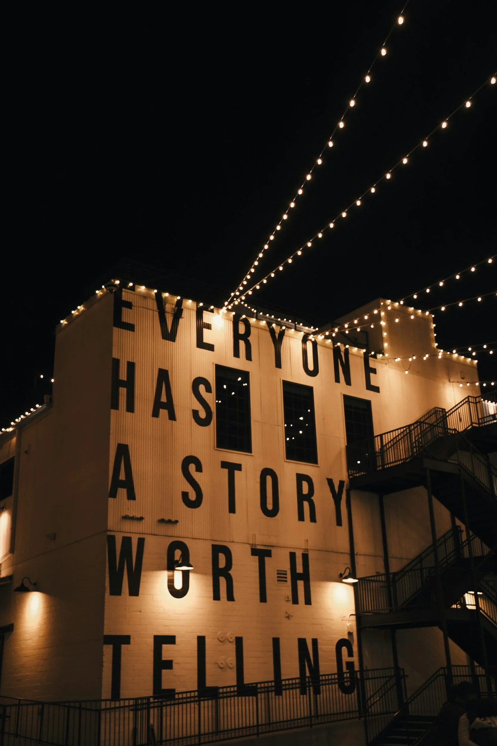 Nighttime view of a building with a large sign on it that reads 'EVERYONE HAS A STORY WORTH TELLING.' String lights are hung above the building, creating a warm glow against the dark sky.