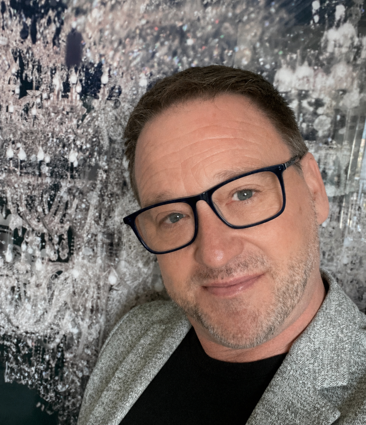 A man with short brown hair, gray beard, wearing glasses and a gray blazer, smiling at the camera, with a sparkling, abstract, silver and black background.