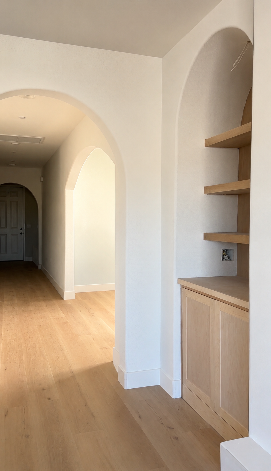 Empty room with light wood flooring, white walls, and arched doorways. Built-in wooden shelving unit with open shelves and closed cabinet.Interior renovation featuring white oak flooring, arched details, and warm neutral finishes by Well, Lived Home.
