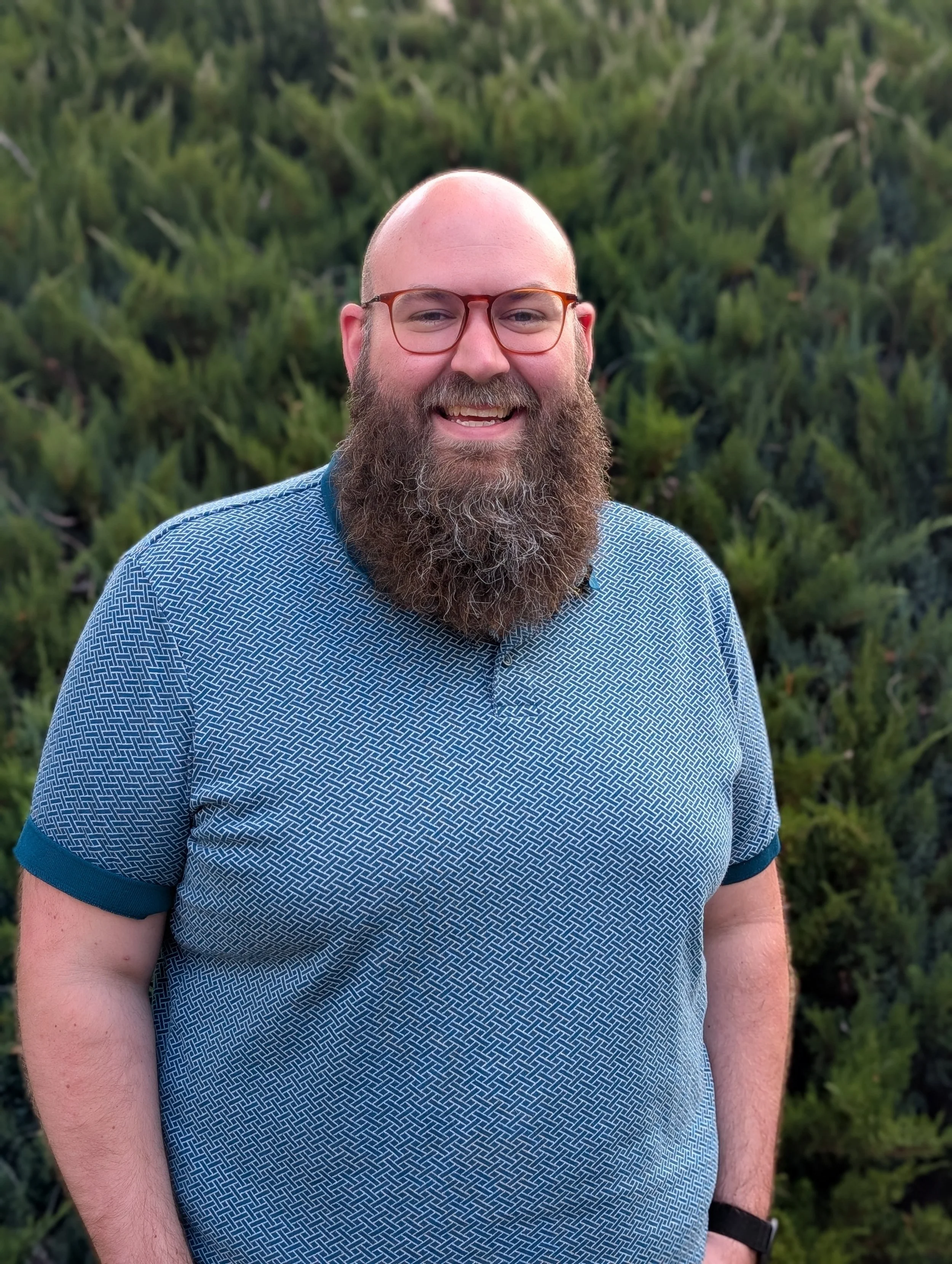 Close-up of a smiling man with a beard, glasses, wearing a patterned blue shirt, standing outdoors with green foliage in the background.