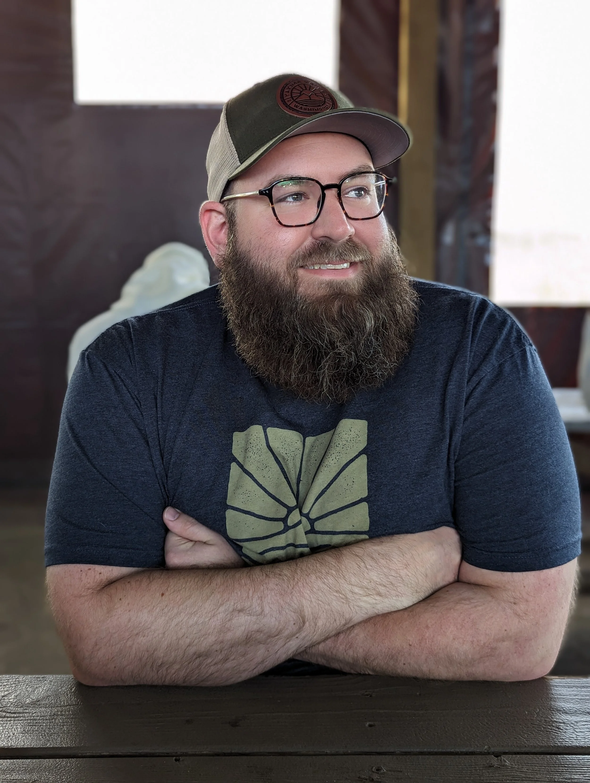A man with glasses and a beard, wearing a gray cap and a black t-shirt, is sitting with his arms crossed on a dark wooden table. The background is blurred.
