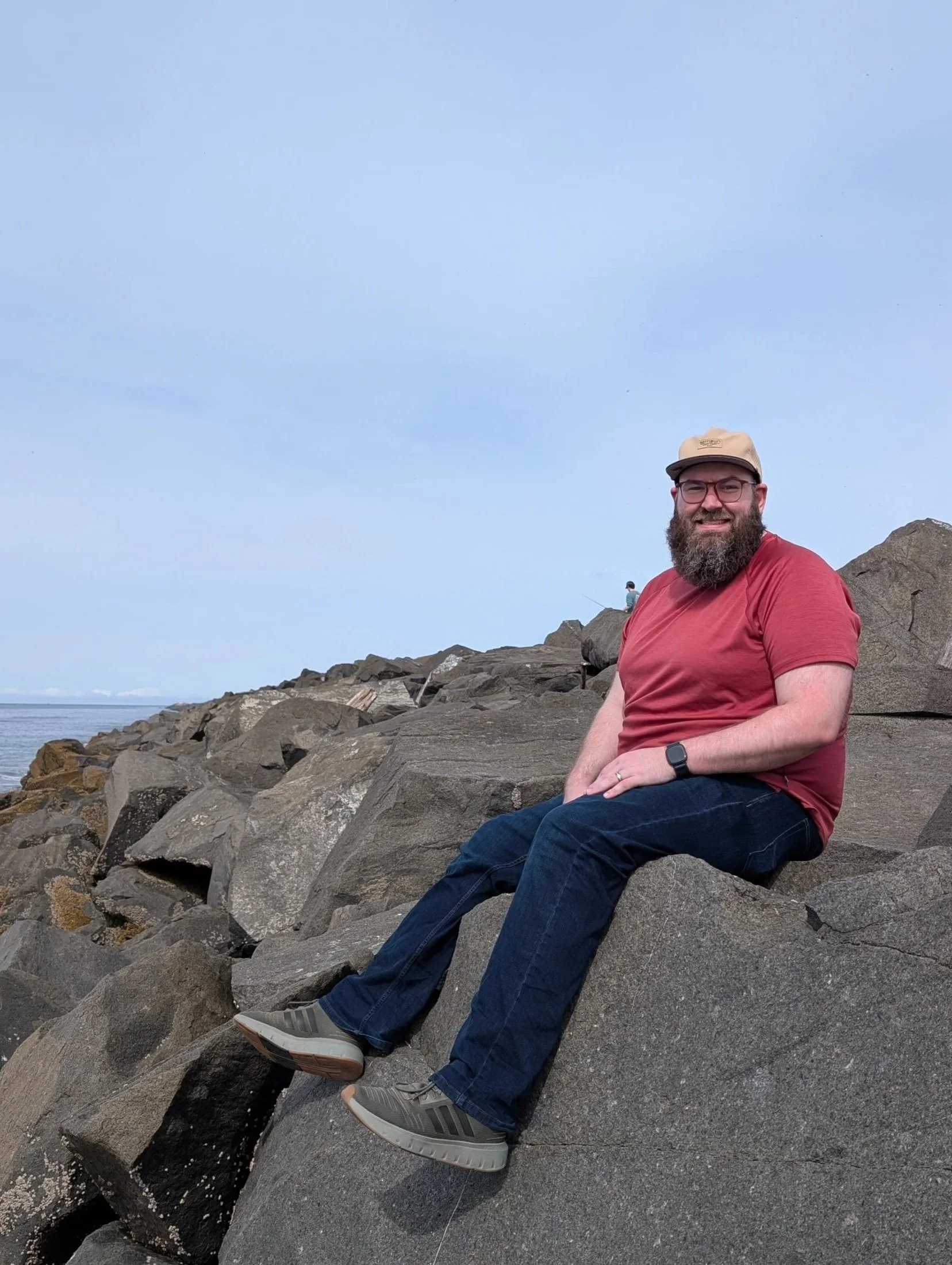 A man with a beard, glasses, red shirt, and beige cap sitting on rocks by the water at the beach, with another person in the background fishing.