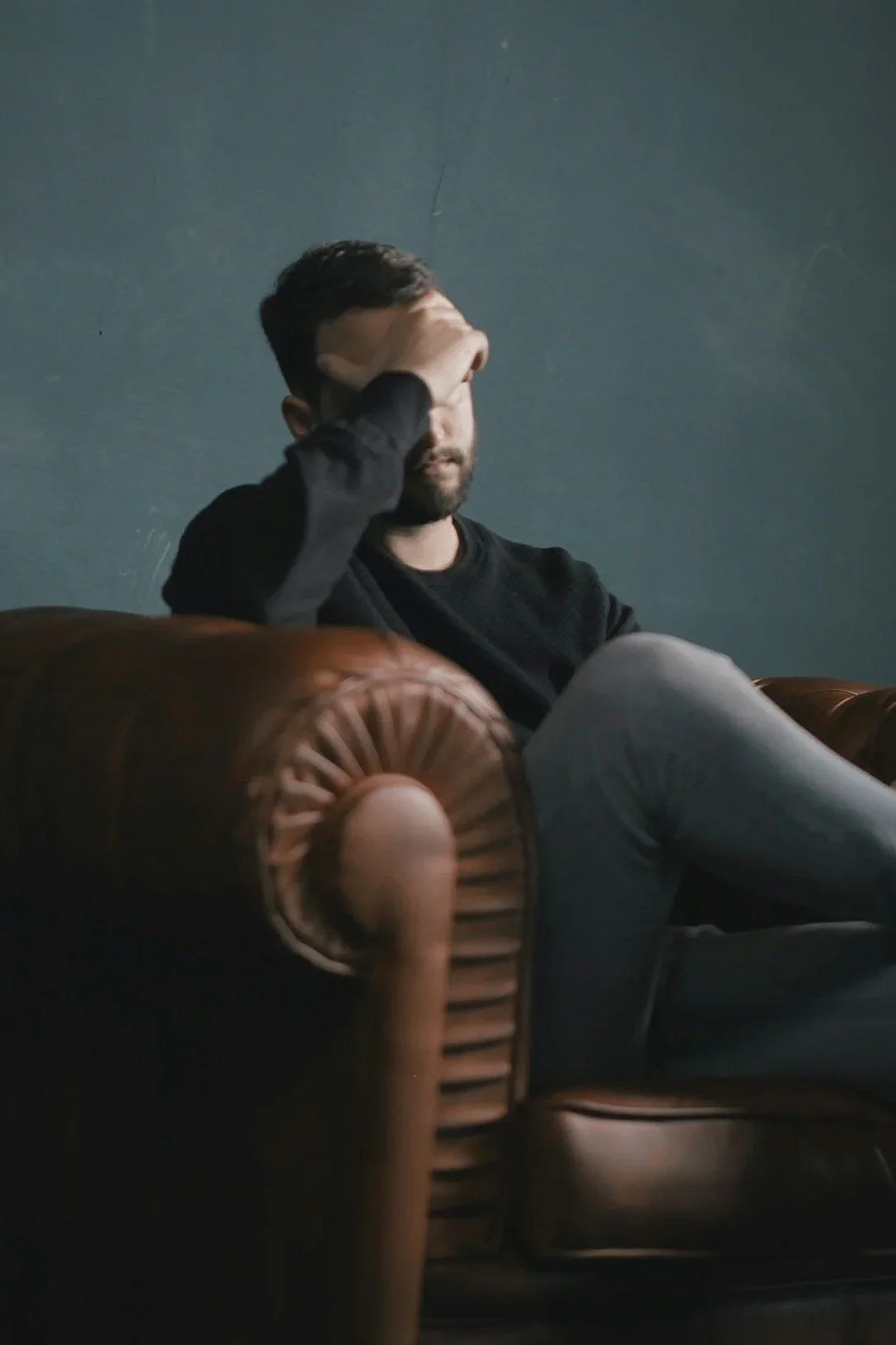 A man sitting on a leather sofa, resting his head in his hand, with a dark wall behind him.