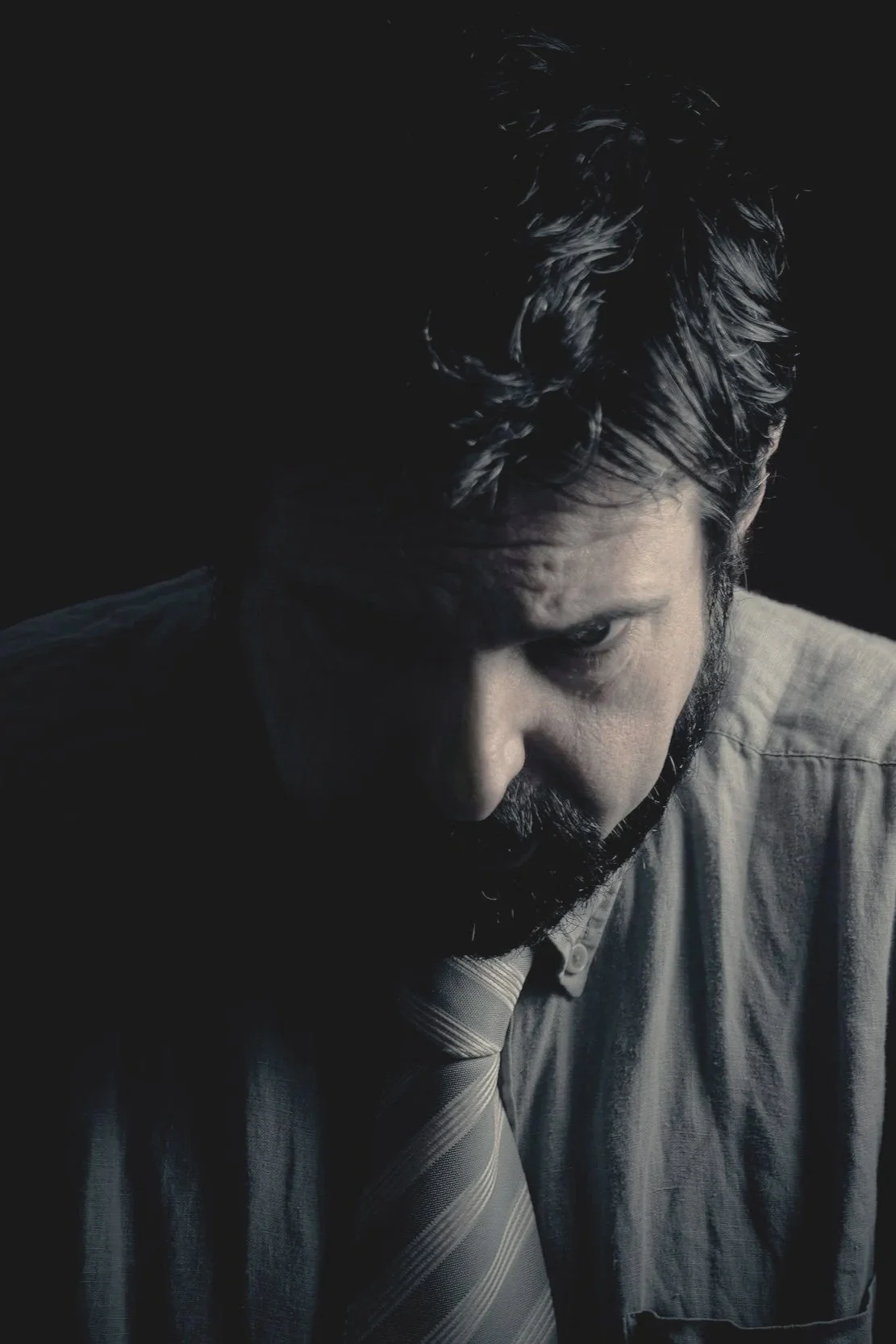 A man with dark hair and a beard looking intensely into the camera, dressed in a shirt and tie, against a dark background.