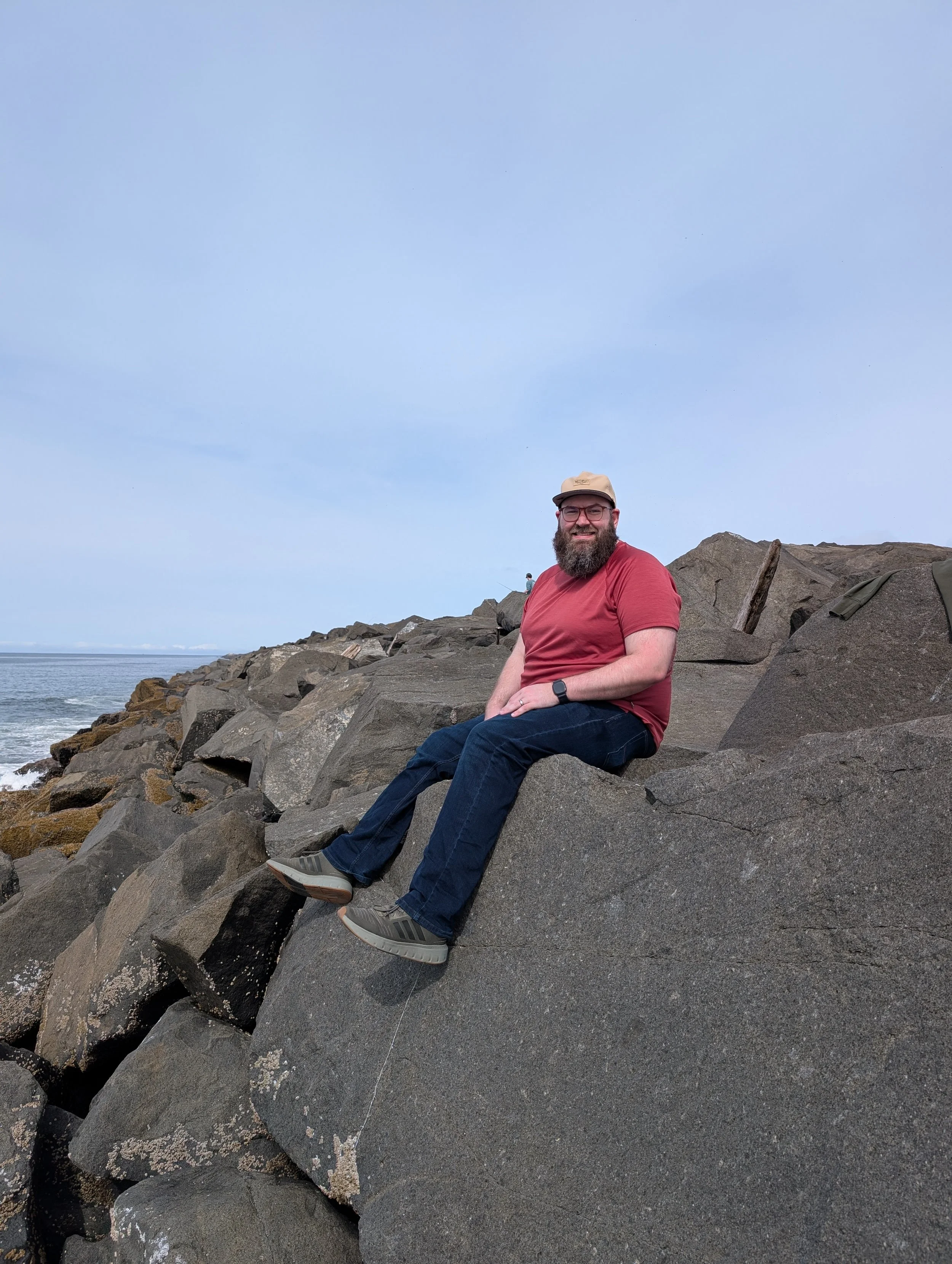 A man with glasses, a beard, and a beige cap sitting on large rocks near the ocean, smiling at the camera.