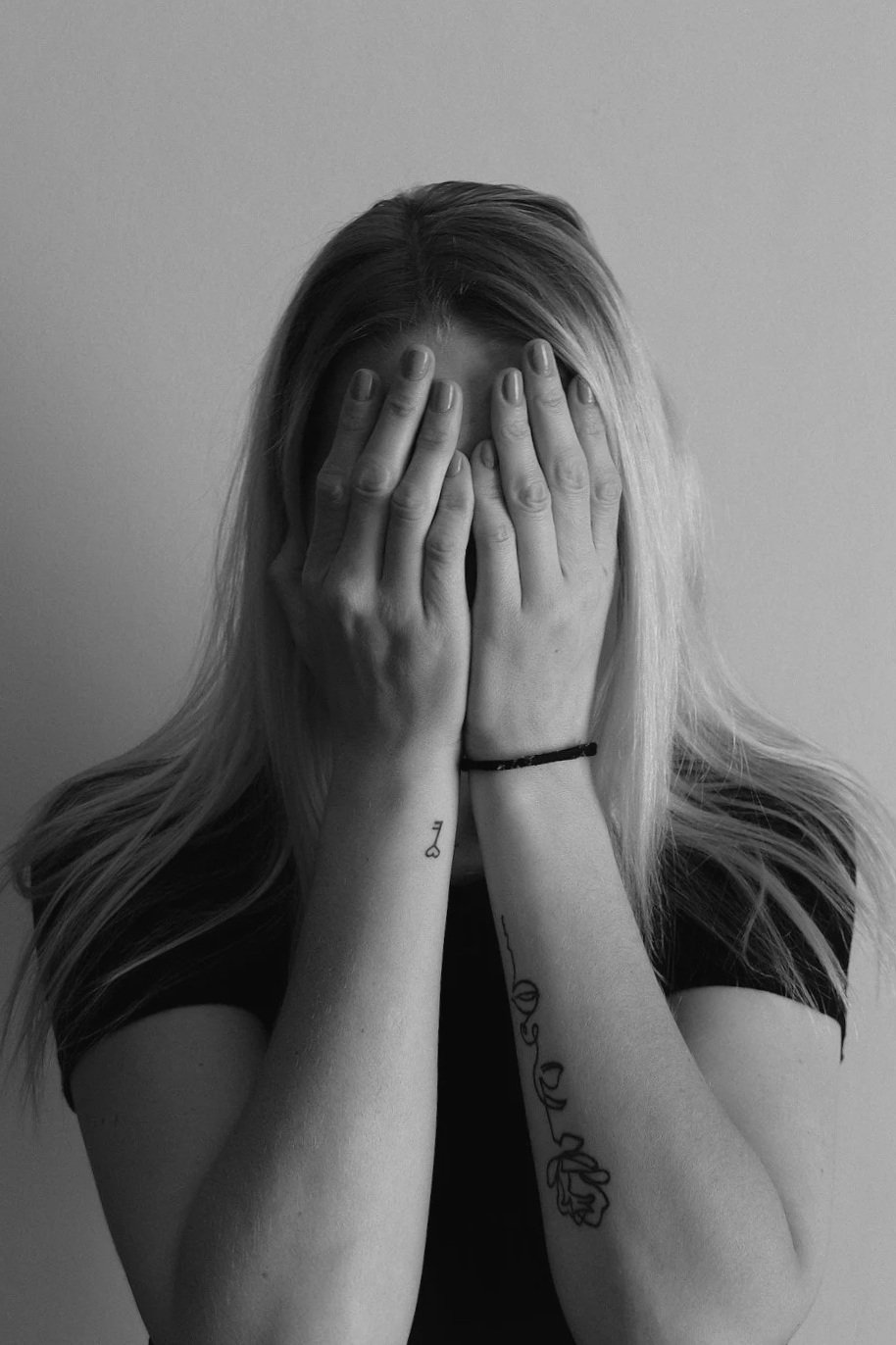 A woman with long, light-colored hair covering her face with both hands, tattoos on her forearms, and a black bracelet on her wrist, standing against a plain background.
