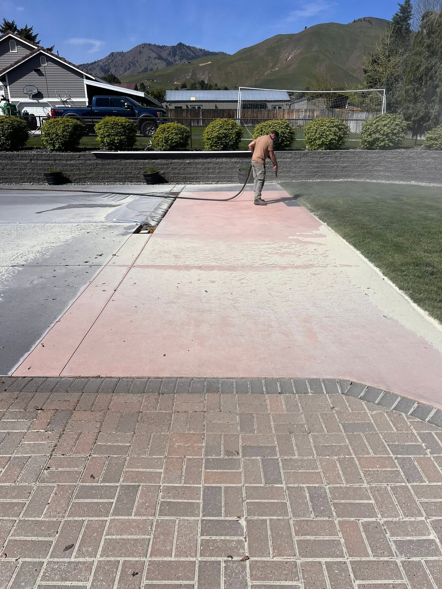 Man cleaning a colorful painted sidewalk with a spray hose in a backyard with green lawn, bushes, a volleyball net, and mountains in the background.