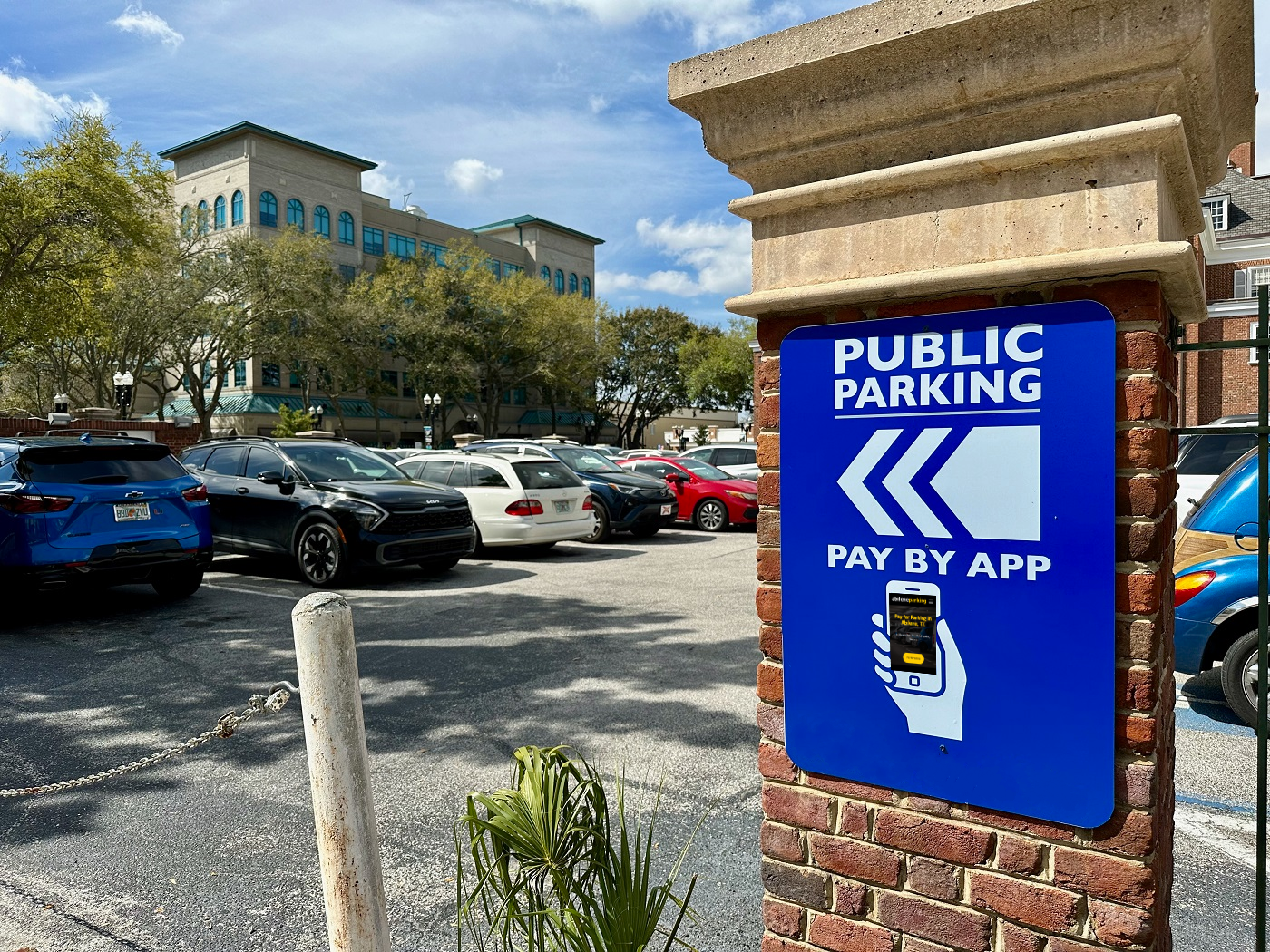Parking lot with multiple parked cars, a blue and white sign indicating public parking and pay by app, and trees in the background under a partly cloudy sky.