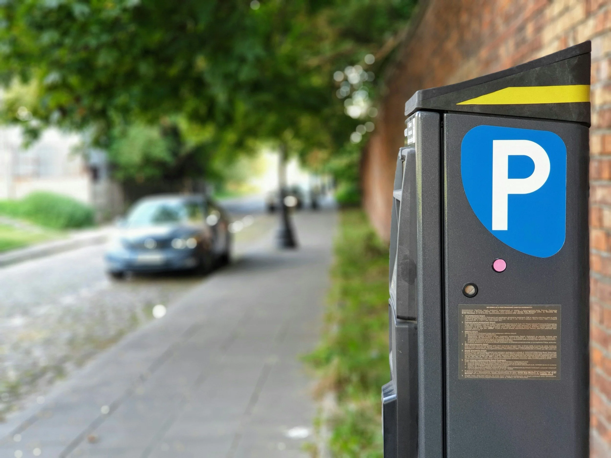 Parking meter on a sidewalk with a blue parking sign, street and blurred cars and trees in the background.