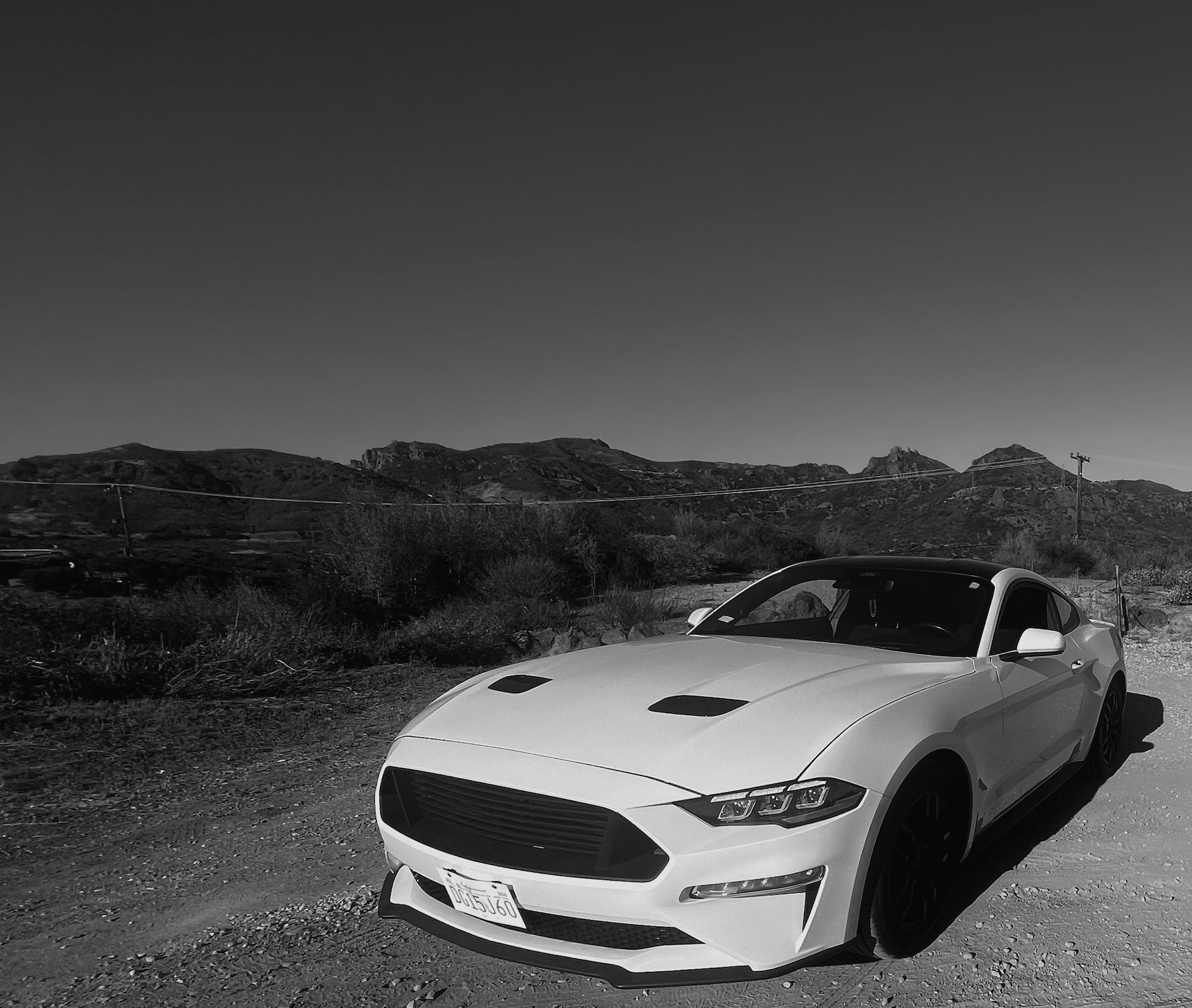A white sports car parked on a dirt road in front of a mountainous landscape under a clear sky.