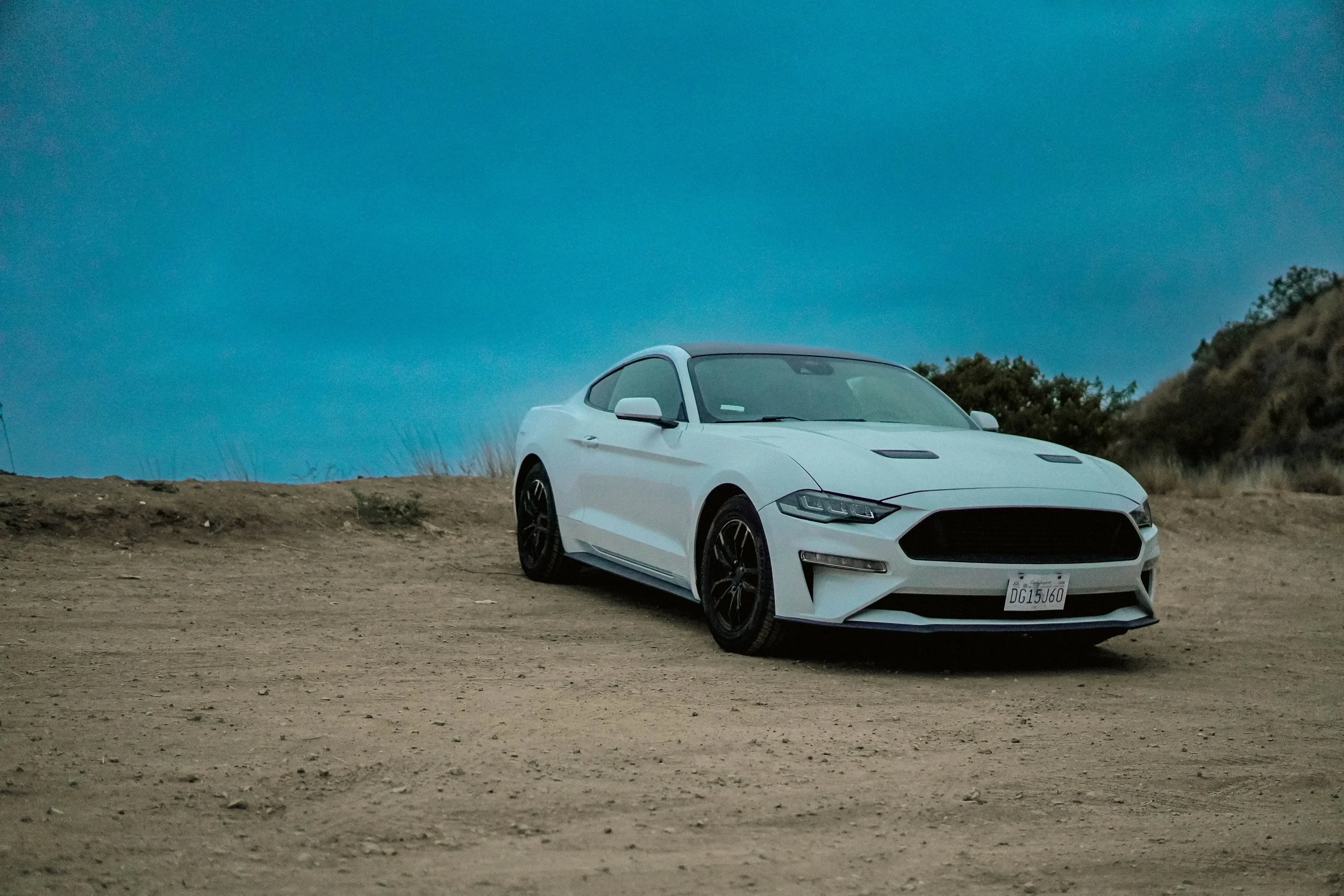 A white sports car parked on a dirt hill under a blue sky, with some vegetation on the right side.