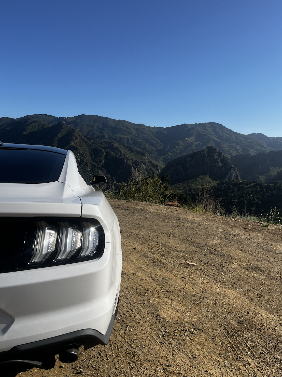 Partial view of a white sports car parked on a dirt road, with green mountainous terrain and a clear blue sky in the background.