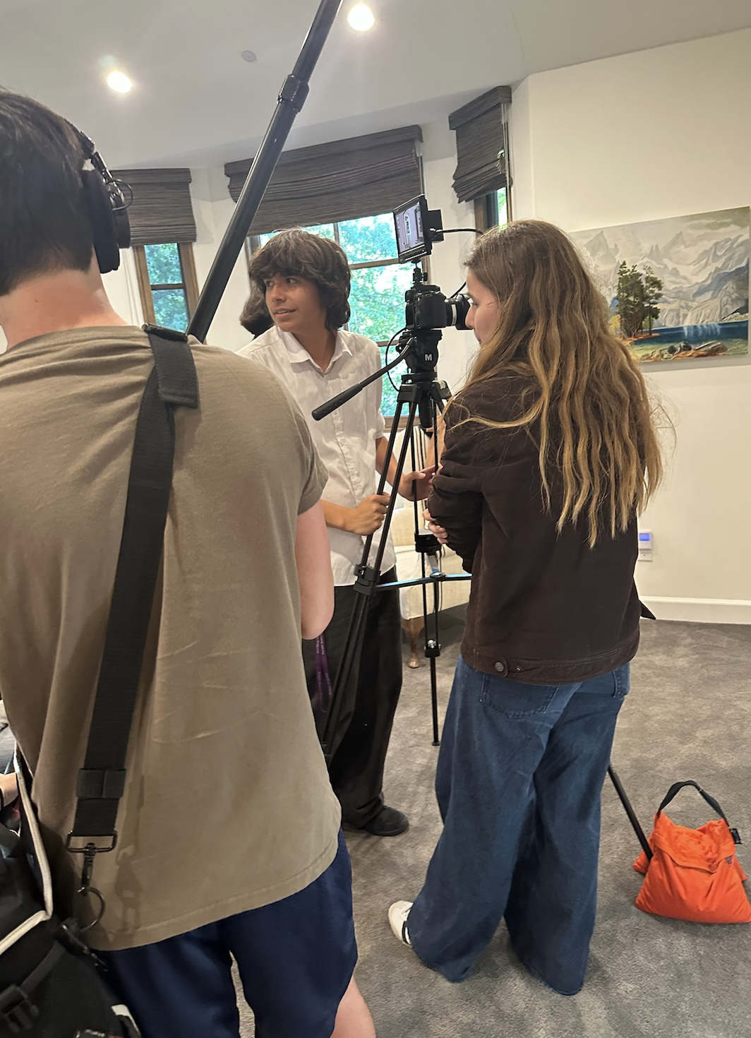 A young man in a white shirt is being interviewed by a woman with long hair and a dark jacket in a room with large windows and artwork on the walls. Two crew members are filming the interview with a camera and a boom microphone.