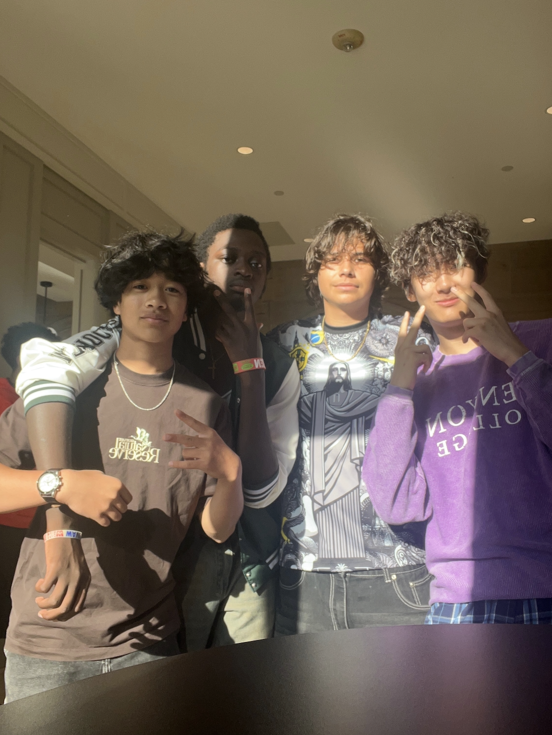 Four young men posing together indoors, two flashing peace signs, one with hand on chin, all casually dressed with jewelry and styled hair.