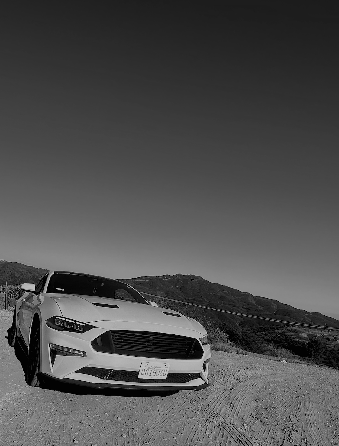 A white sports car parked on a dirt road with mountains in the background, image in black and white.