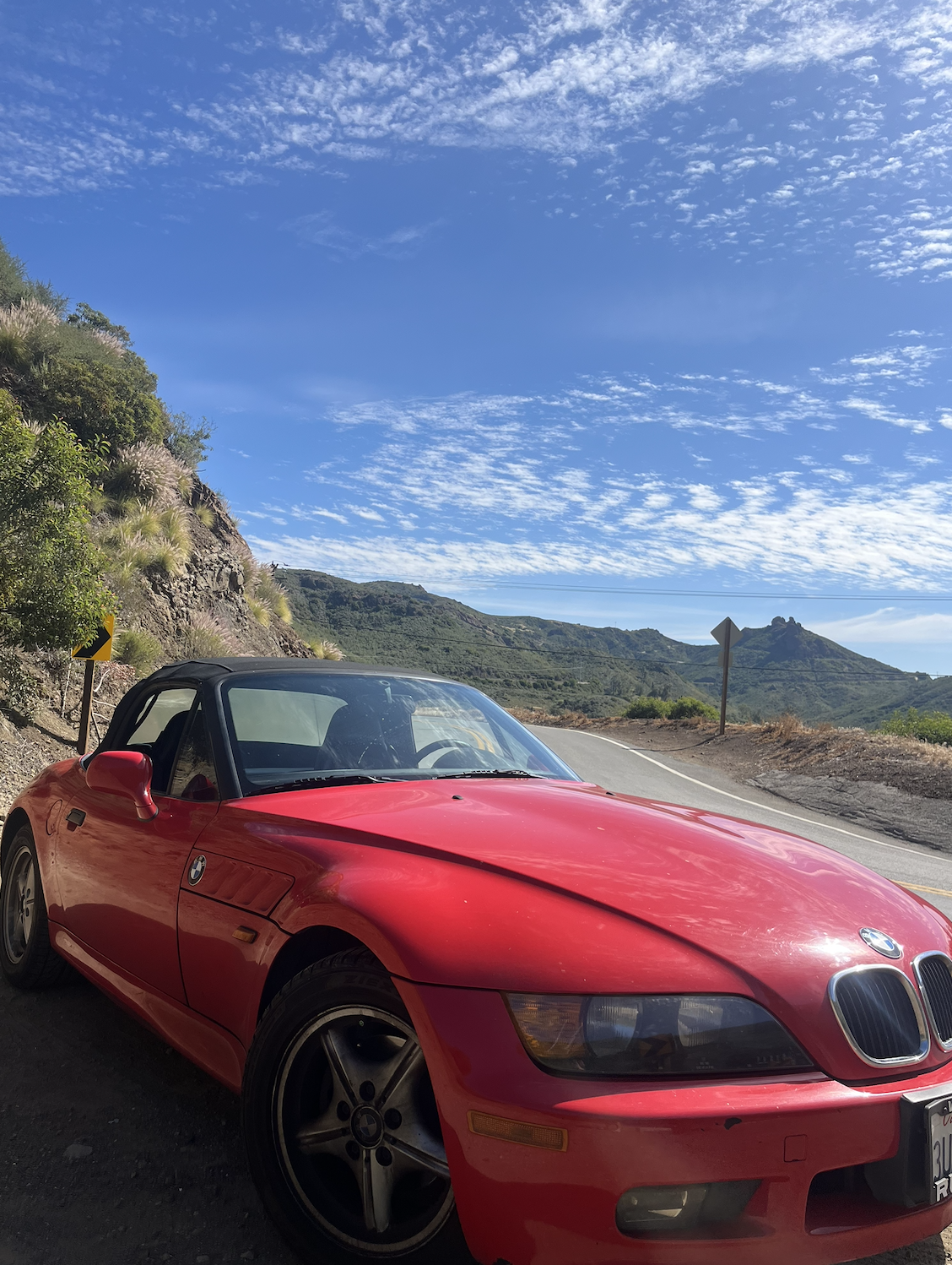 A red BMW convertible parked on a mountain road with a scenic background of green hills and a blue sky with wispy clouds.
