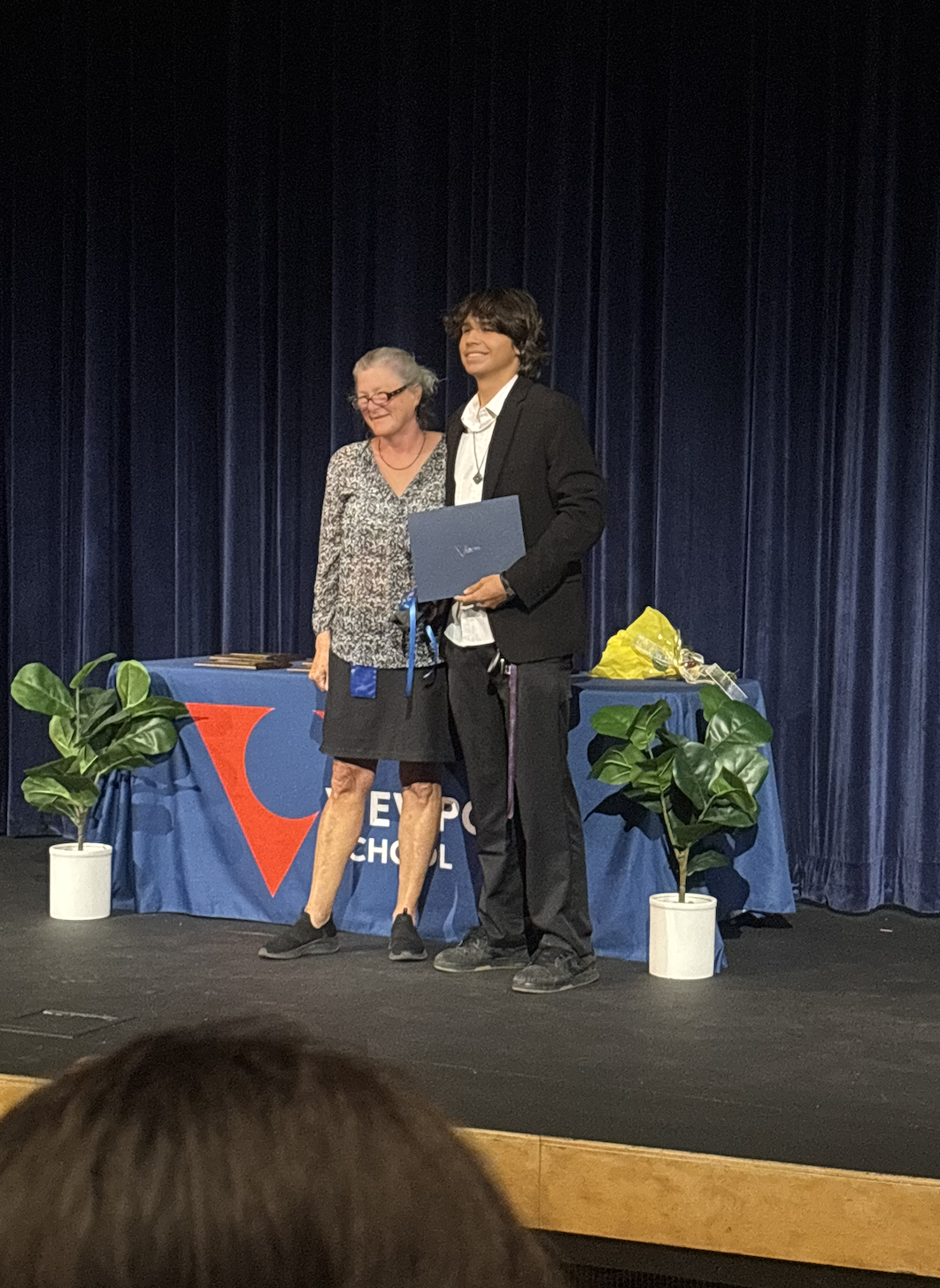Young man in black blazer receiving diploma from older woman on stage at a graduation ceremony, with a table holding flowers and awards behind them.