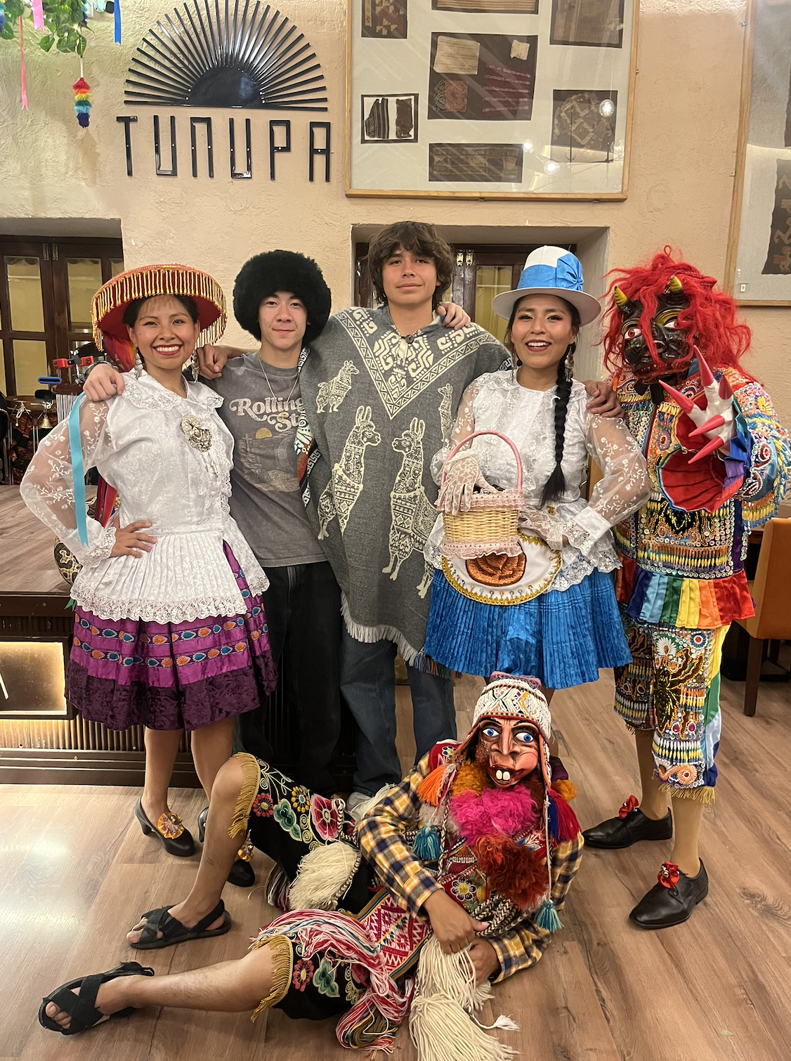 Group of five people in colorful traditional and costume outfits, with one person in a rainbow costume and mask, posing indoors at a restaurant with sign 'TUMUPA' on the wall.
